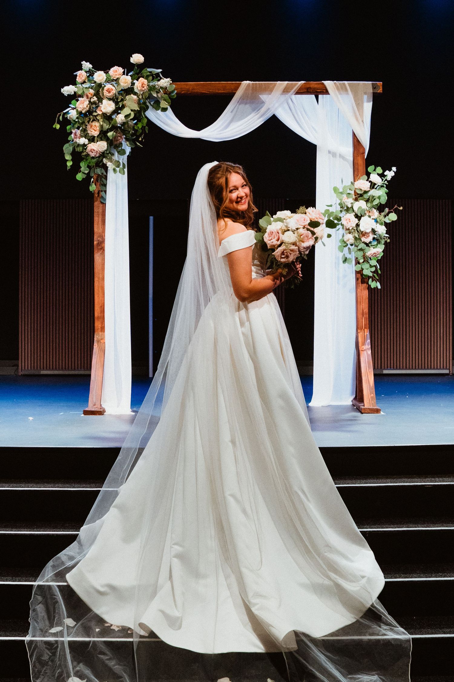 Radiant bride in white gown with veil holding bouquet before a draped floral arch at the altar.