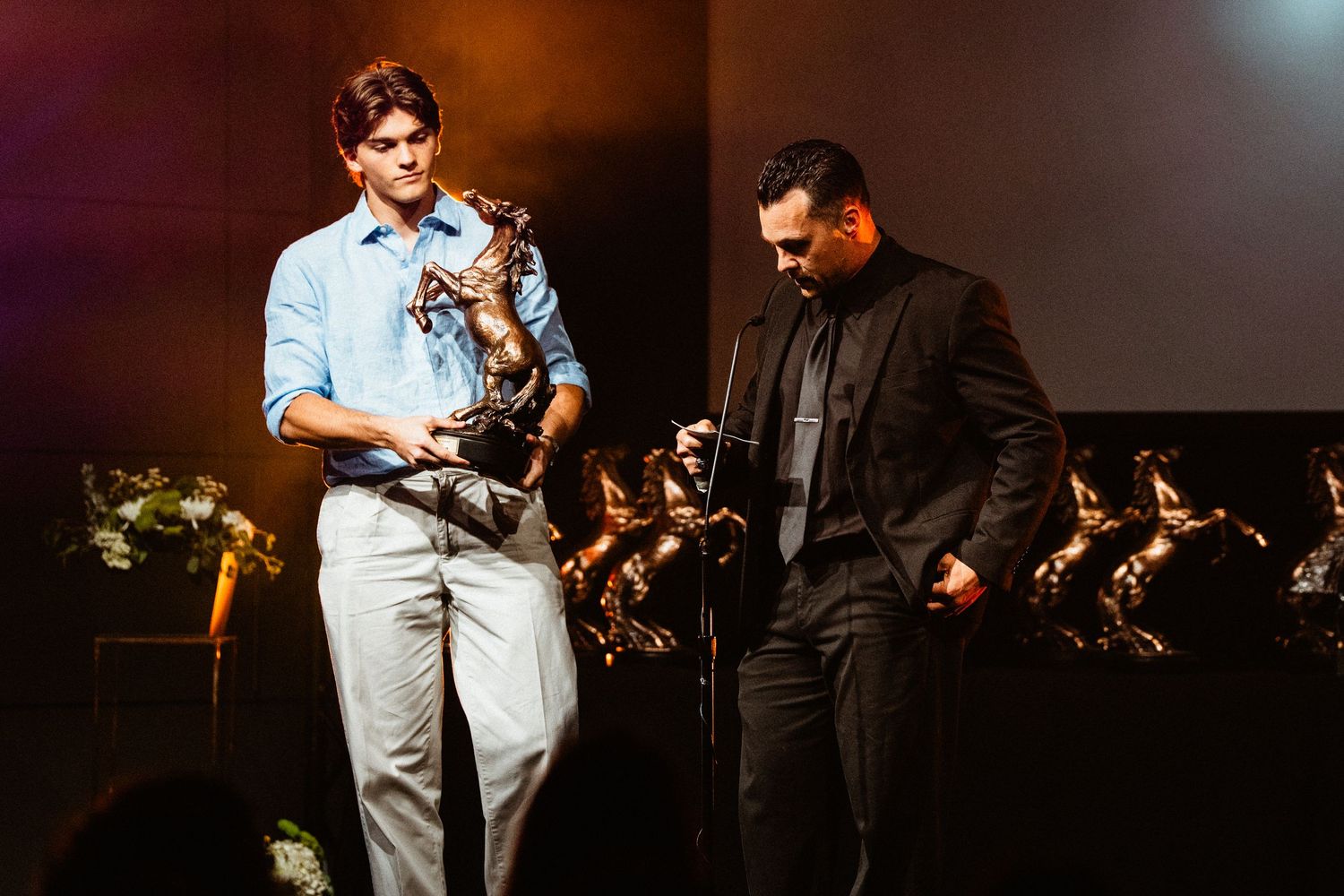 Two men on stage at an awards ceremony, one holding a bronze trophy statue under dramatic lighting.