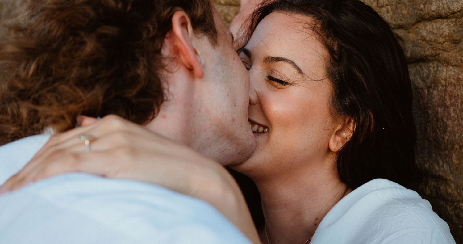 A romantic couple sharing a tender kiss outdoors, with the woman smiling blissfully against a rustic stone wall.