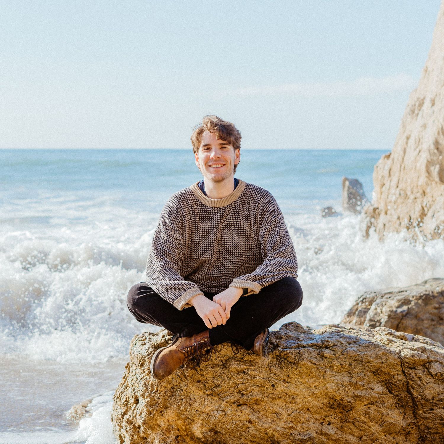 Young man sitting cross-legged on a rock with ocean waves crashing behind him.