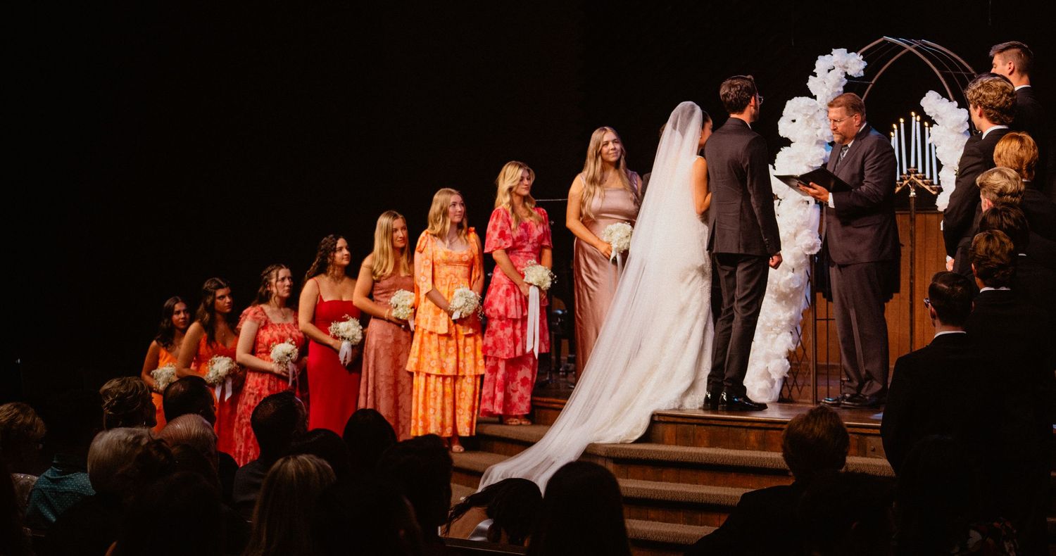 Bride at altar with long veil surrounded by bridesmaids in colorful orange and pink dresses in church.