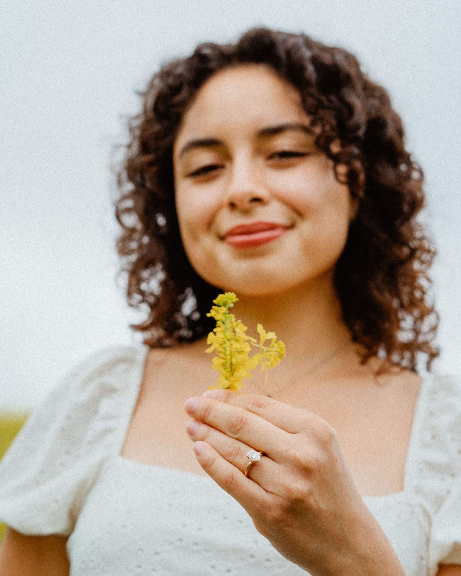 Smiling young woman with curly hair holding a small yellow wildflower in a field.