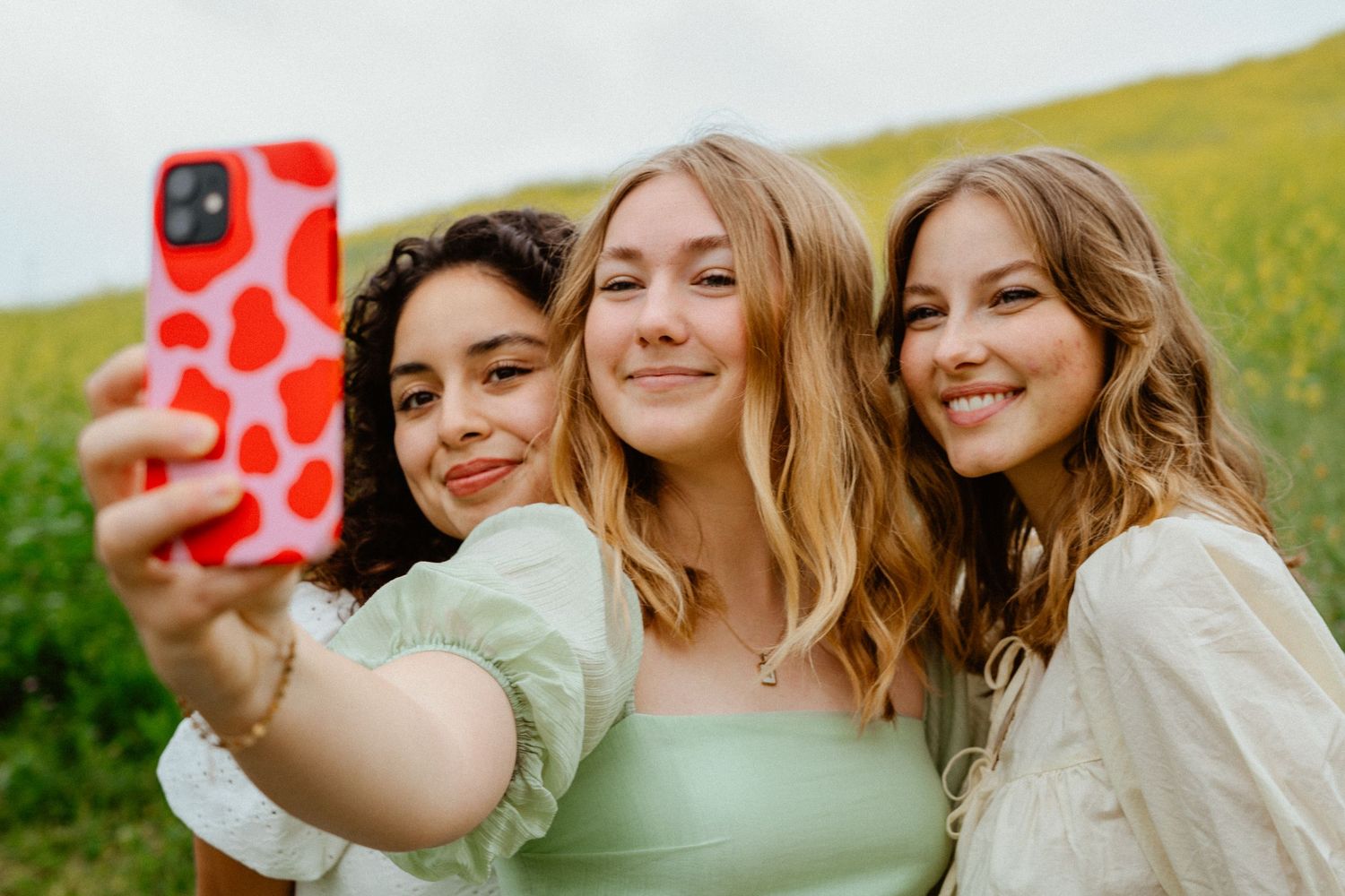 Three smiling young women taking a selfie with a red phone in a yellow flower field outdoors.