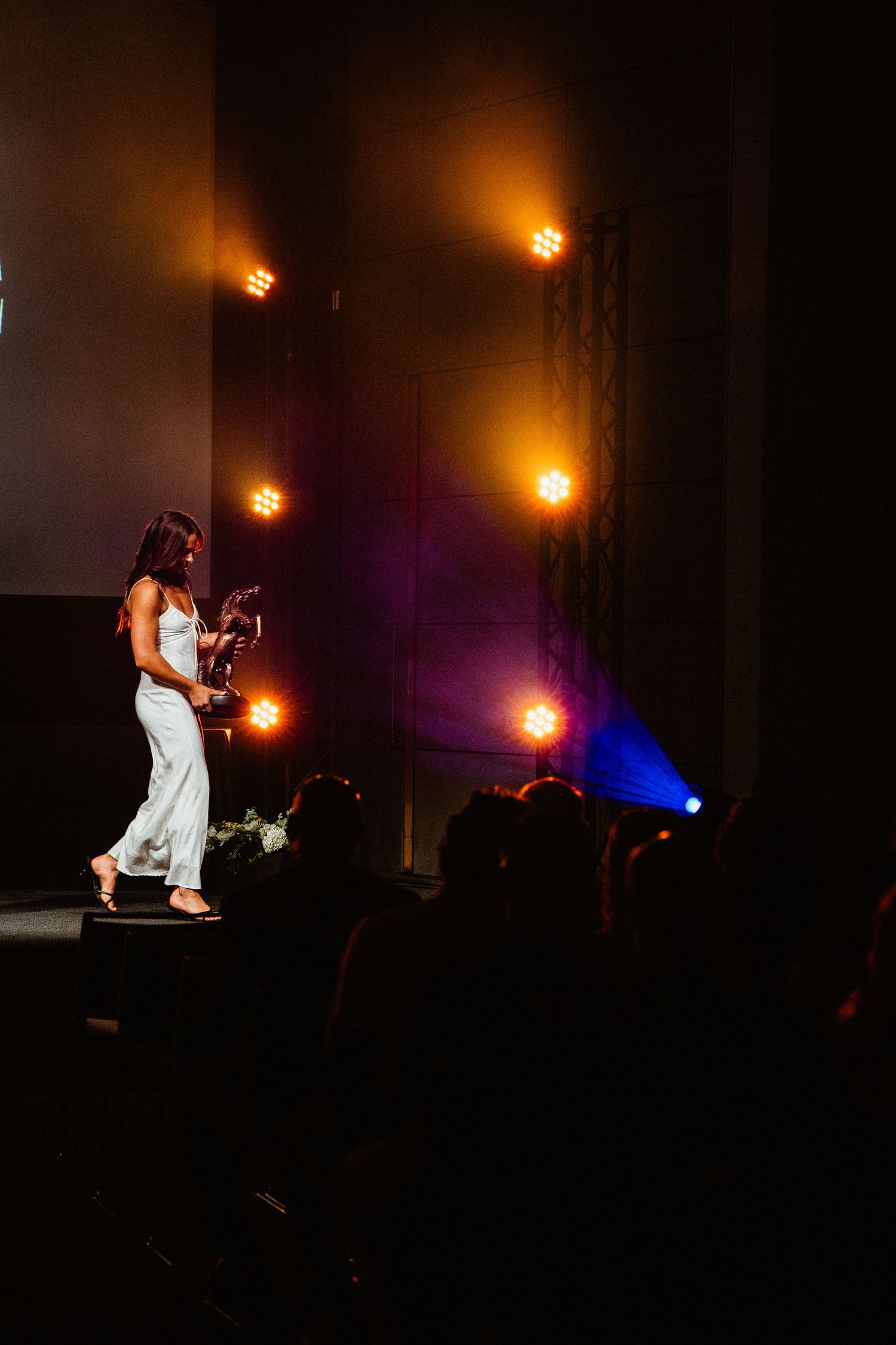 Woman in white outfit accepts award on stage with warm orange stage lighting and audience silhouettes.