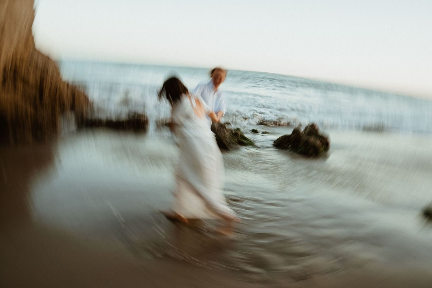 Blurred motion photo of couple in white walking in ocean waves on rocky beach at sunset.