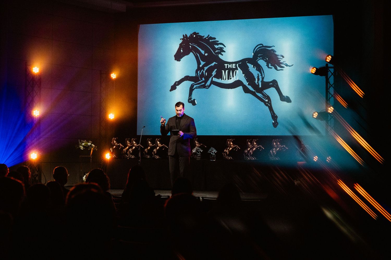 A presenter on stage before a large screen displaying a black horse logo at a dark, dramatic live event.