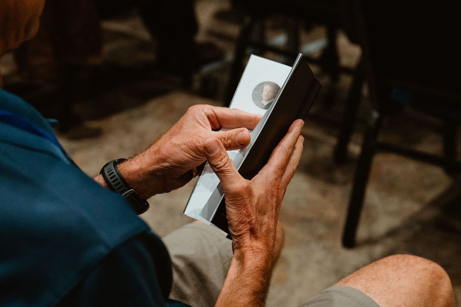 Elderly person holding a smartphone displaying a contact photo, wearing a blue shirt and ring.