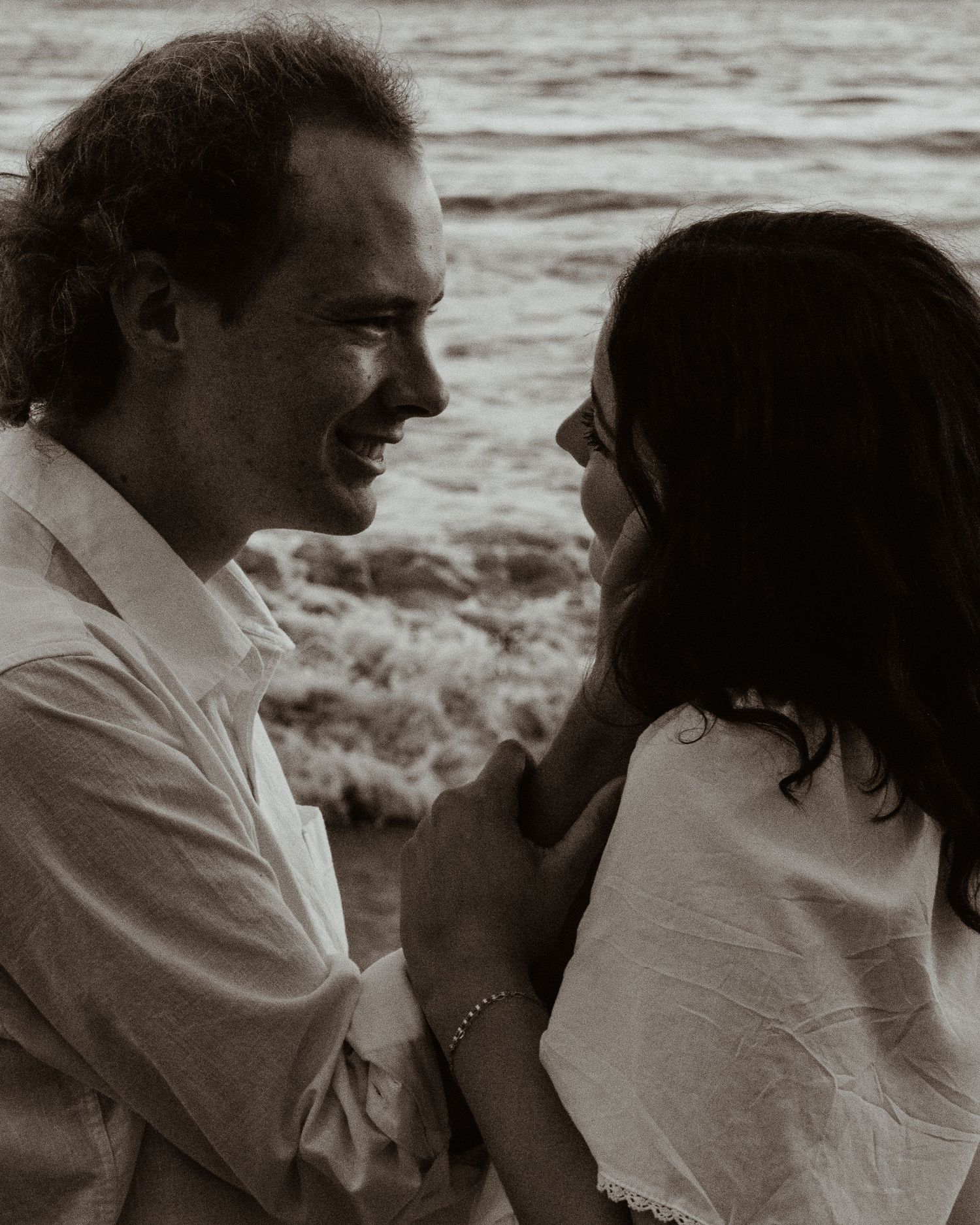 Black and white photo of a smiling couple holding hands by the ocean waves.