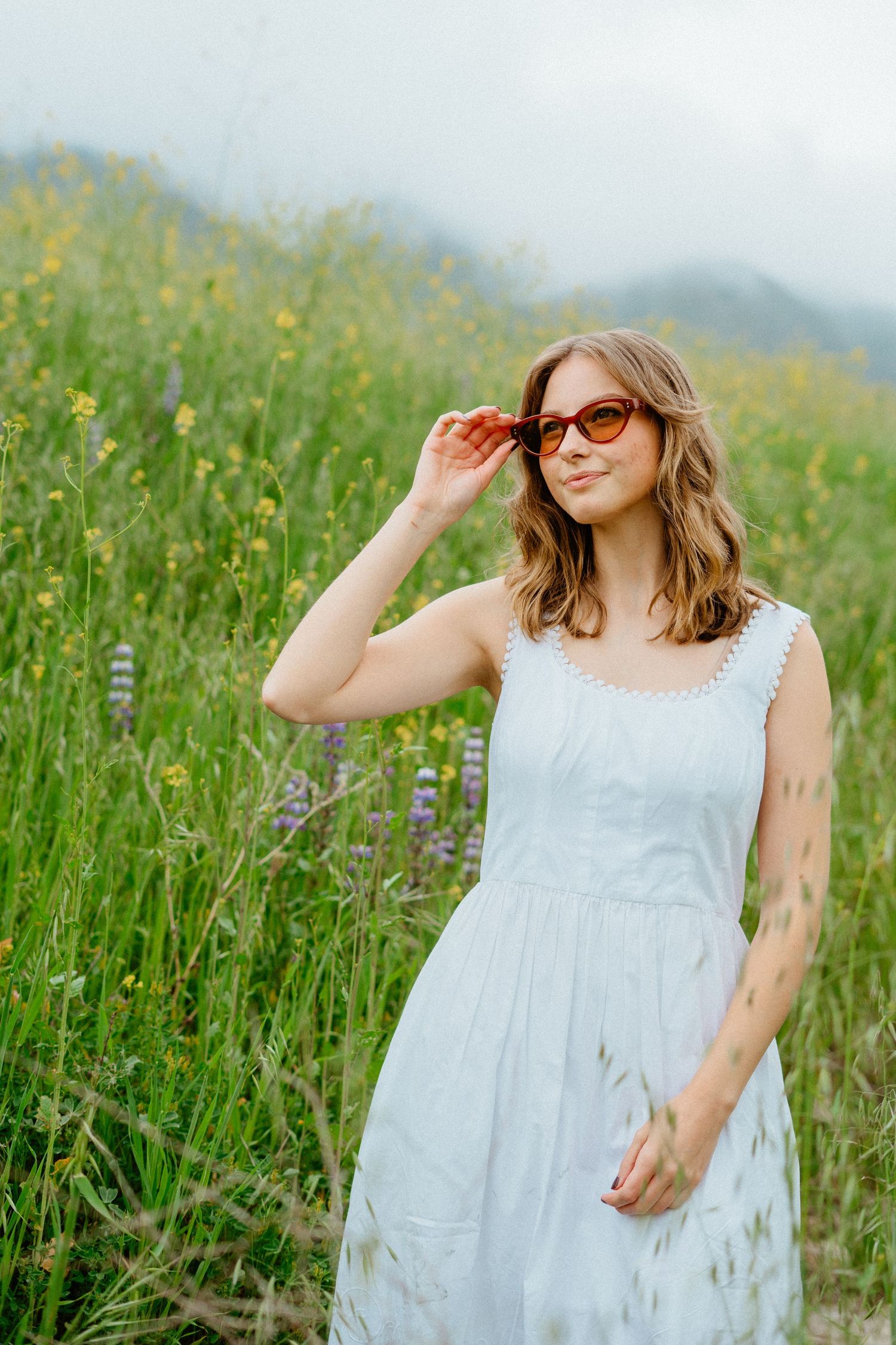 Young woman in white sundress adjusts orange sunglasses while standing in a green wildflower meadow.
