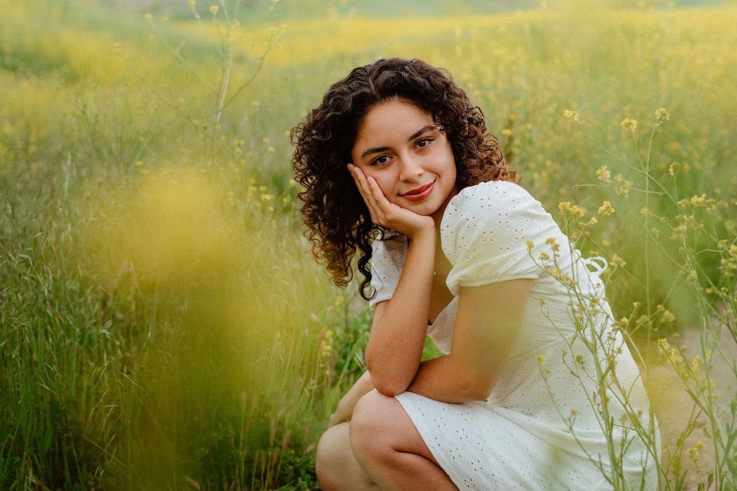 Young woman with curly hair in white dress sitting among yellow wildflowers in a field.