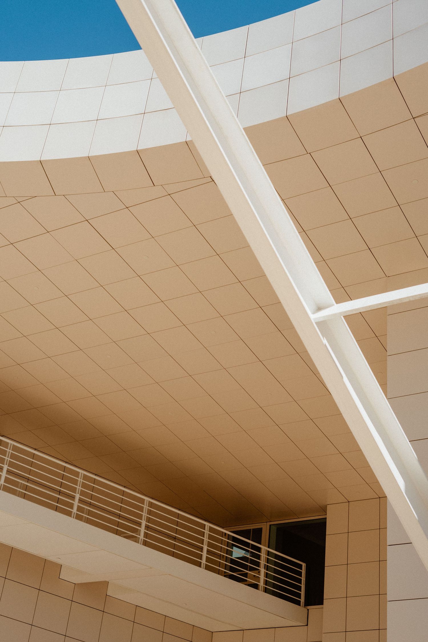 Modern curved building exterior with beige stone cladding, white structural beams, and metal railing balcony against blue sky.