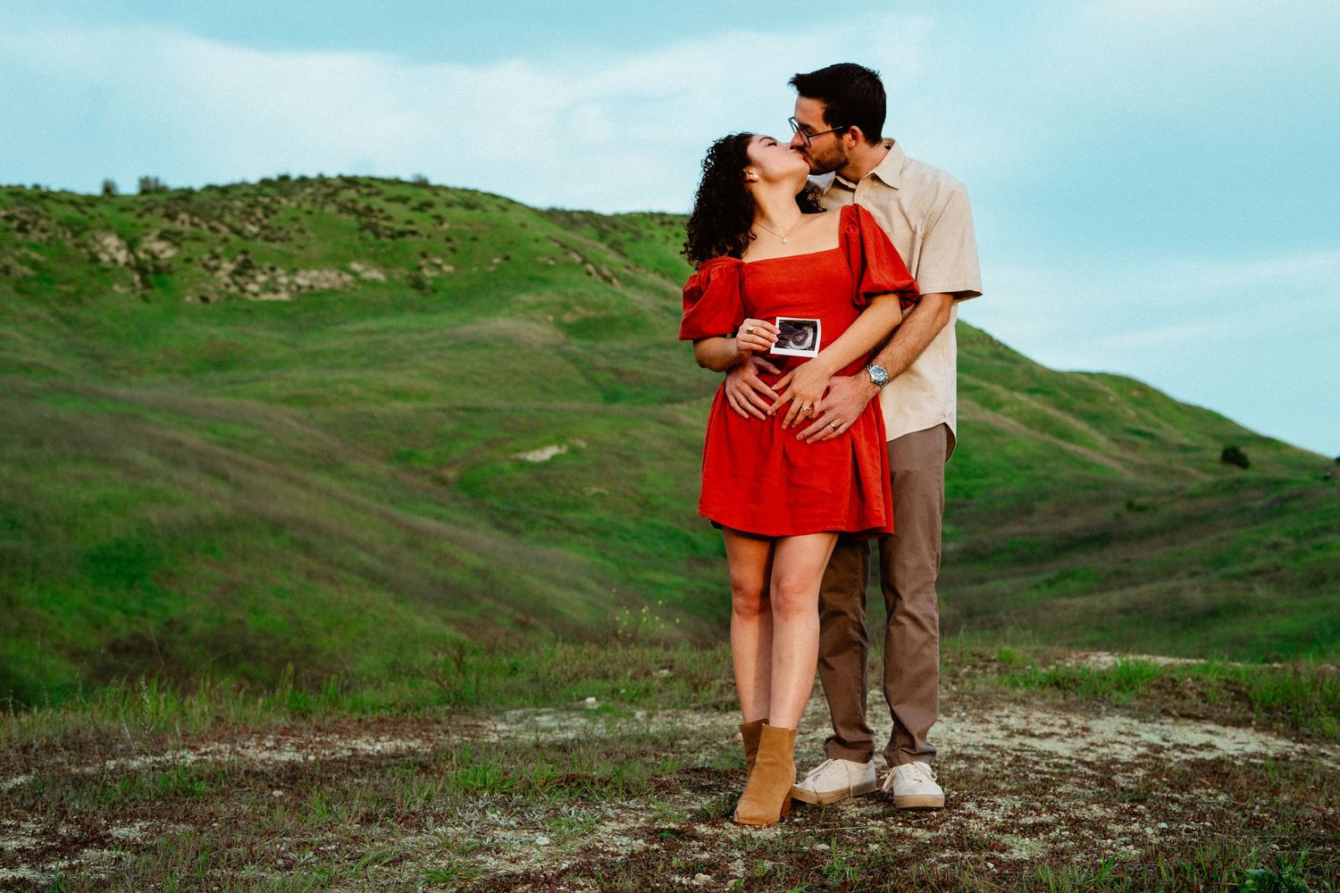 Couple embracing on green hillside, holding ultrasound scan, woman in red dress kissing moment.