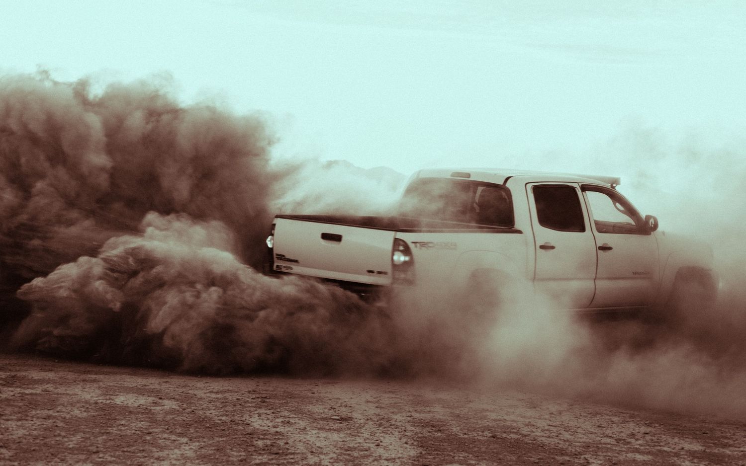 Sepia-toned pickup truck creating large dust cloud while driving across dry desert landscape.