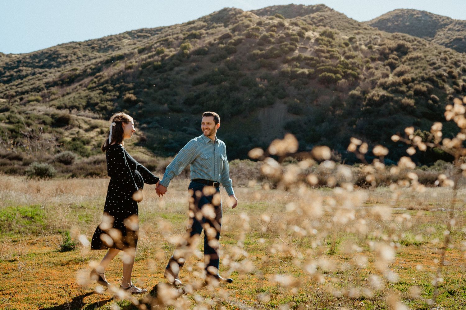 Couple holding hands in sunny field with wildflowers and green hills behind them.