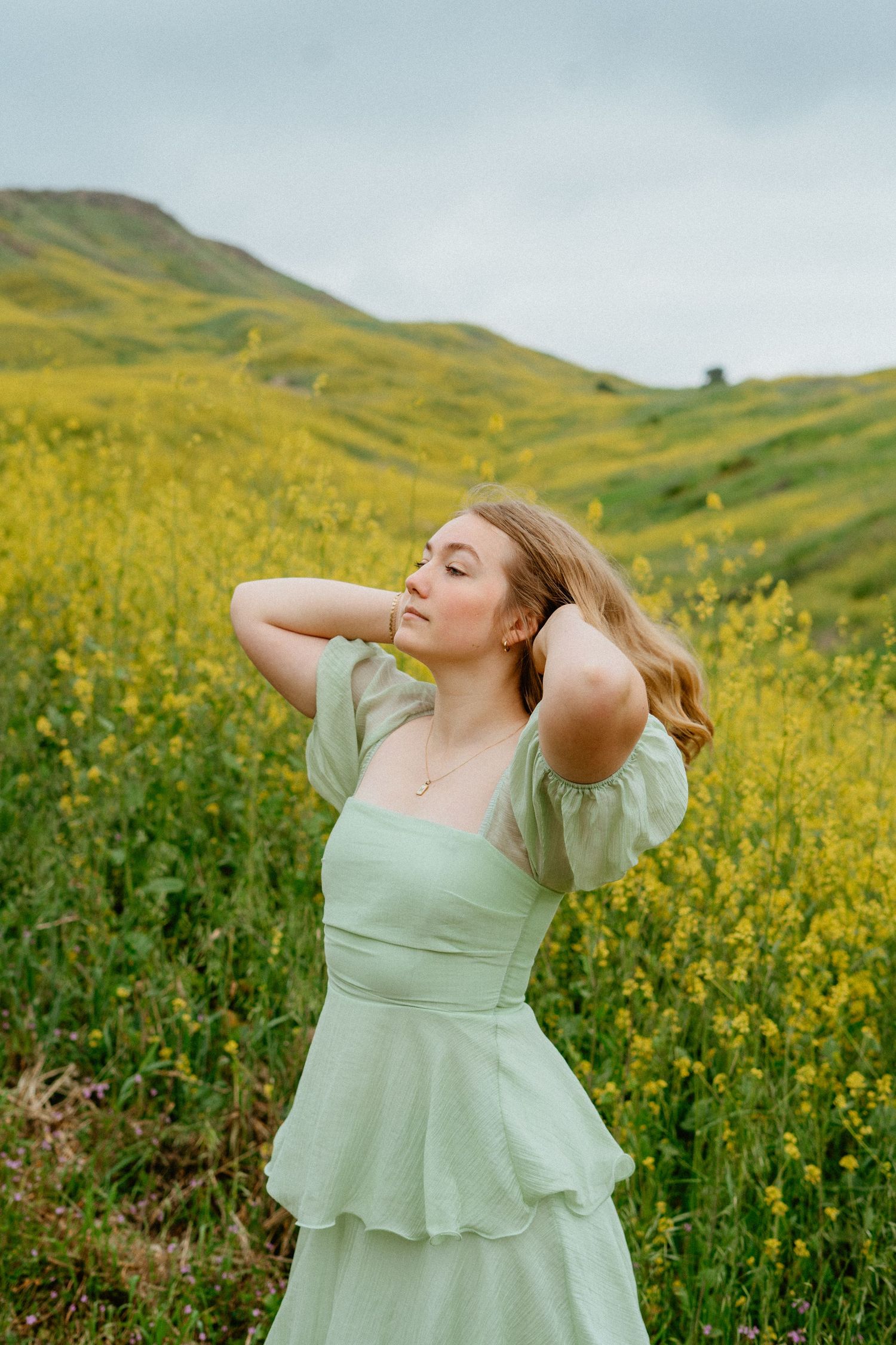 Blonde woman in mint tiered dress posing in yellow flower field with hills.