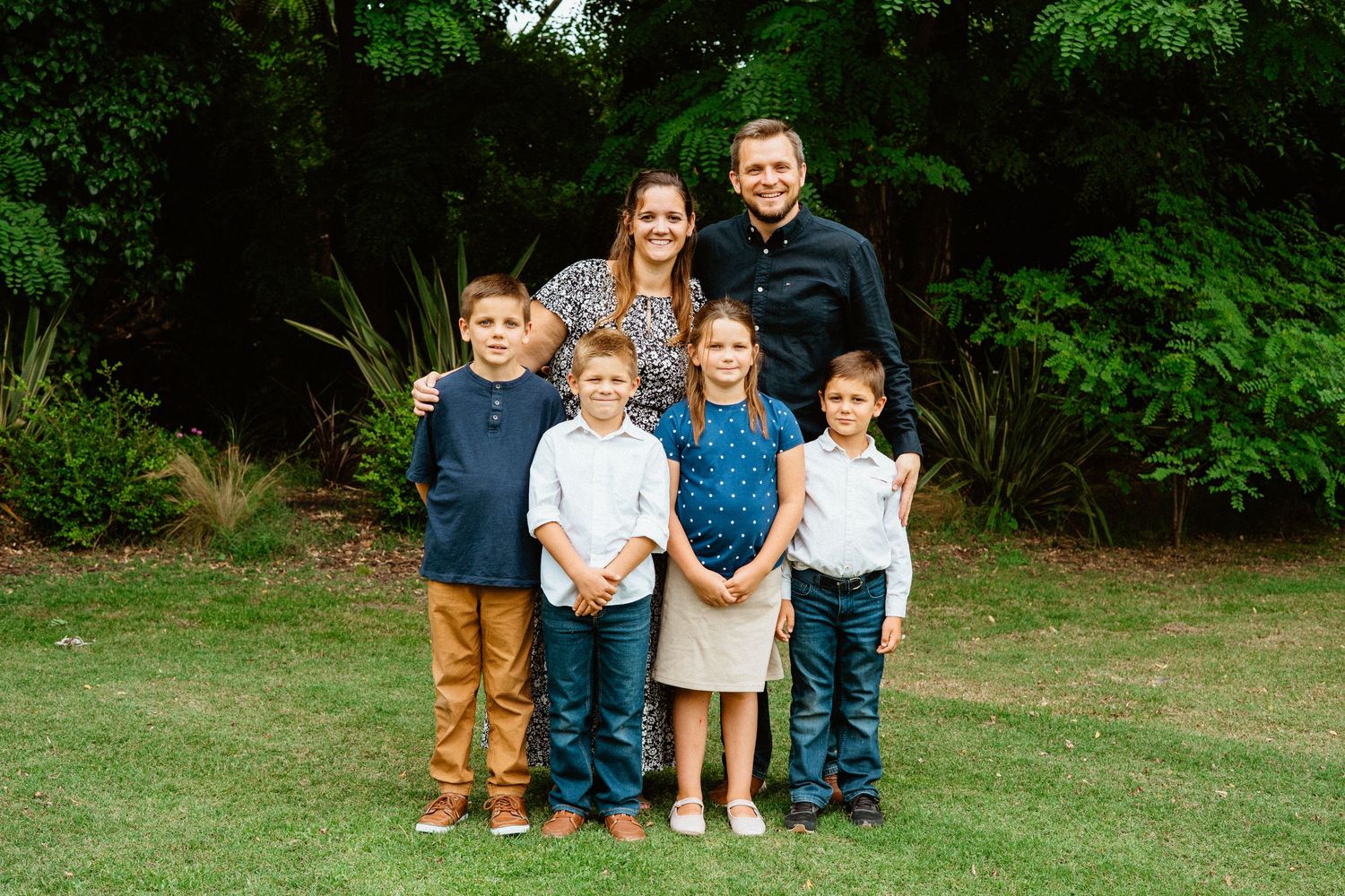 Family of six smiling together on green lawn with trees behind.