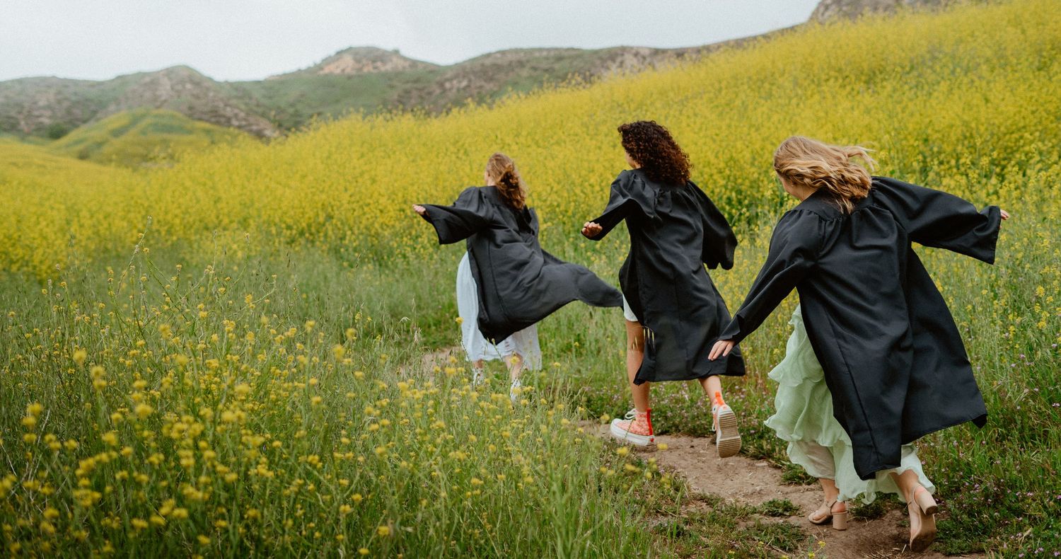 Three women in graduation gowns running through a scenic yellow wildflower field with mountain backdrop.