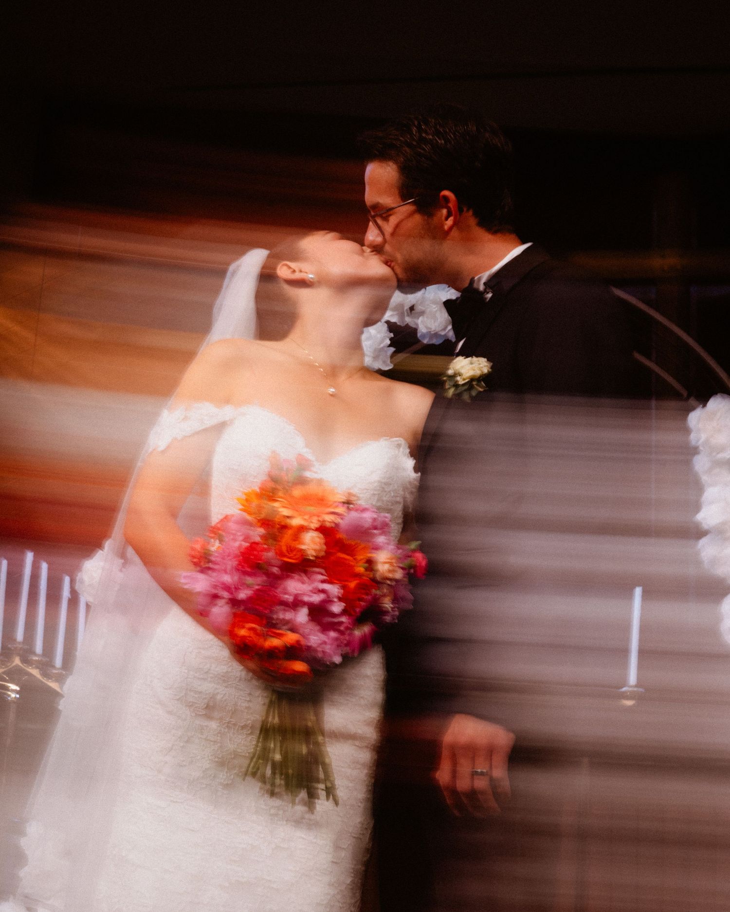 Artistic motion blur photo of bride and groom sharing first kiss, bride holding colorful pink and orange bouquet.
