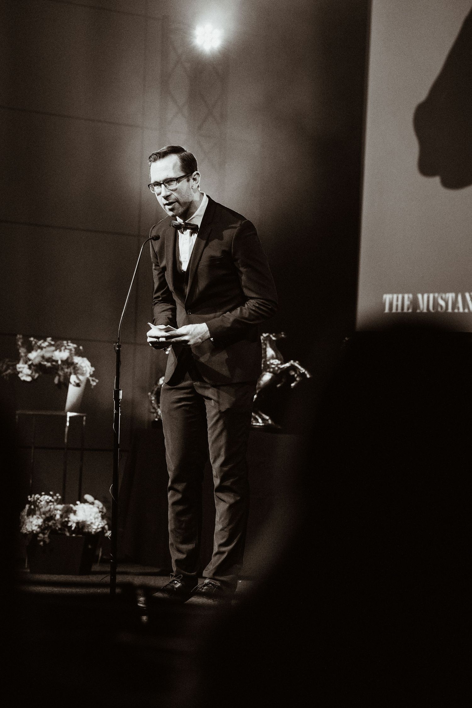 Elegant man in tuxedo speaking at podium during formal awards ceremony on stage.