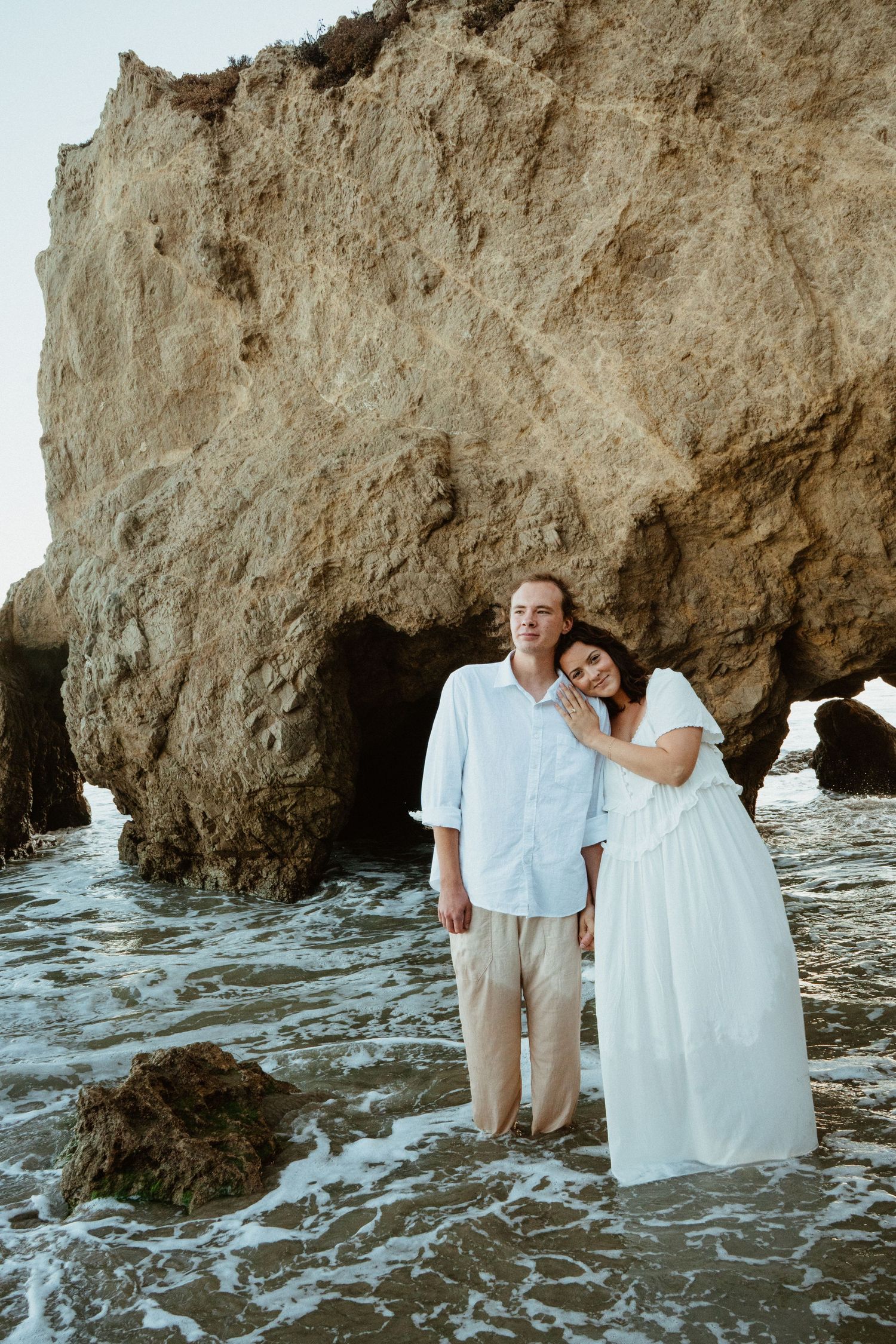 Couple embracing barefoot in ocean waves beside a large rocky cliff on a scenic beach.
