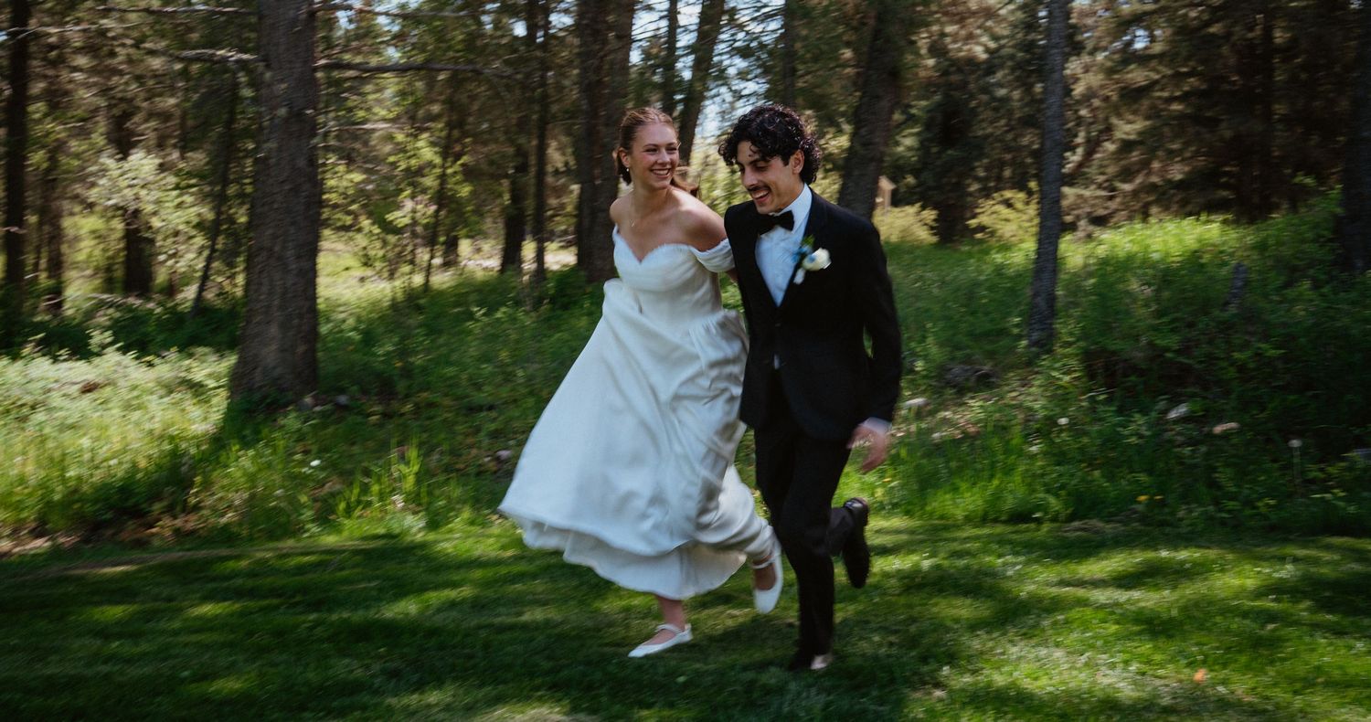 Joyful newlyweds in wedding attire running together on green grass surrounded by tall pine trees outdoors.