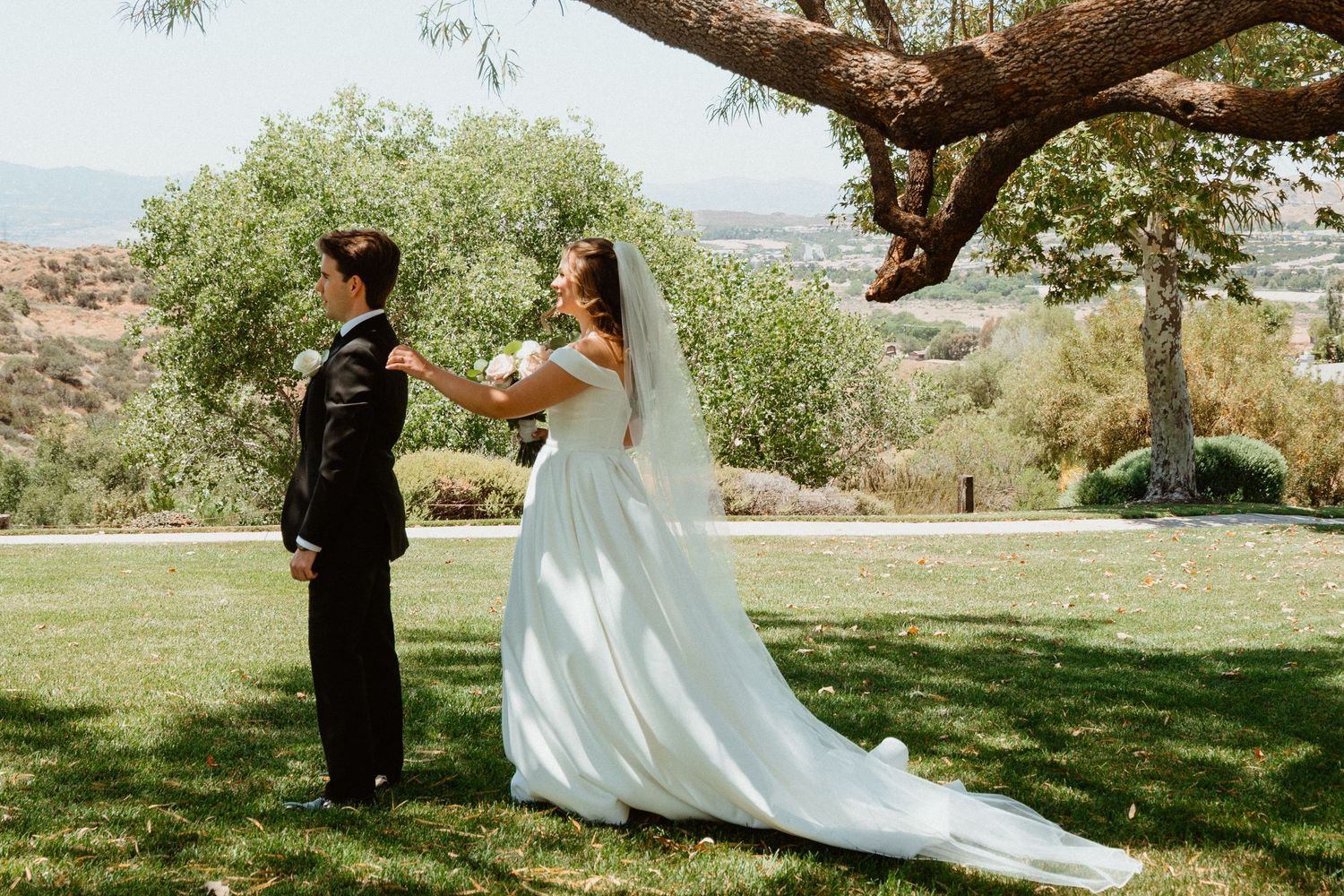 Bride in white gown with veil stands behind groom in black tux outdoors under tree with scenic hills.