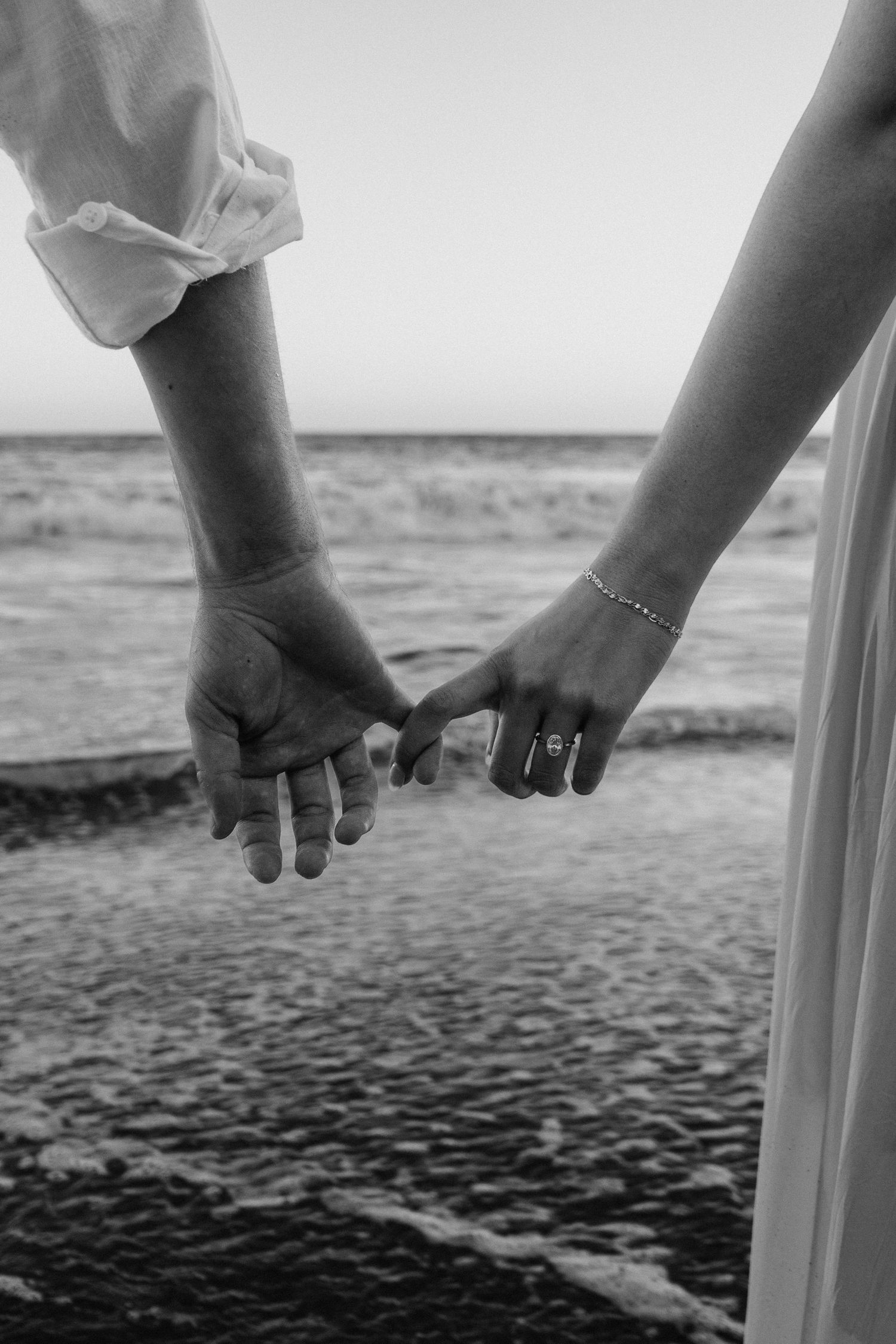 Black and white photo of a couple holding hands on a beach, bride wearing a diamond ring and bracelet.