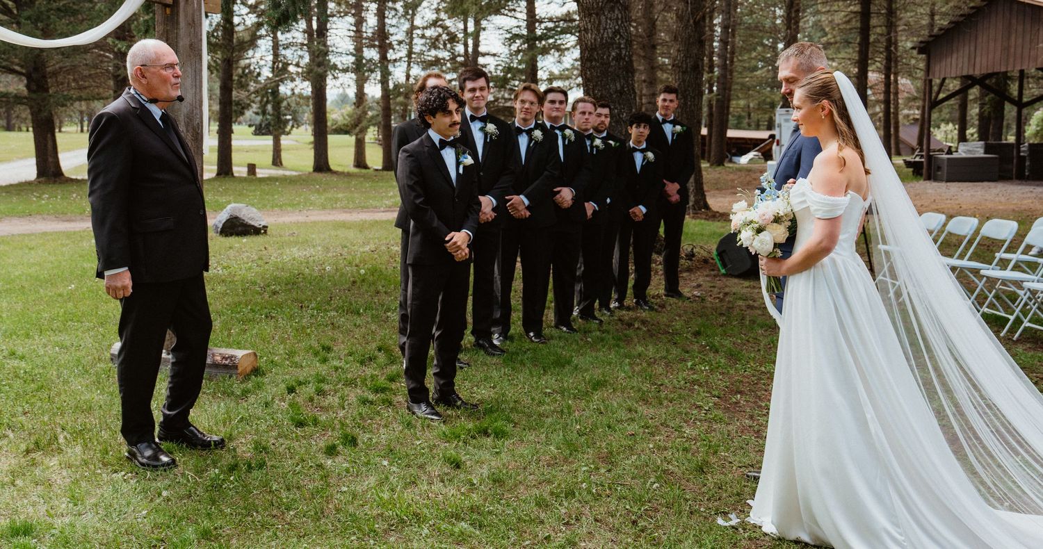 Outdoor wedding ceremony with bride in white gown and veil facing groom and groomsmen in black suits among pine trees.