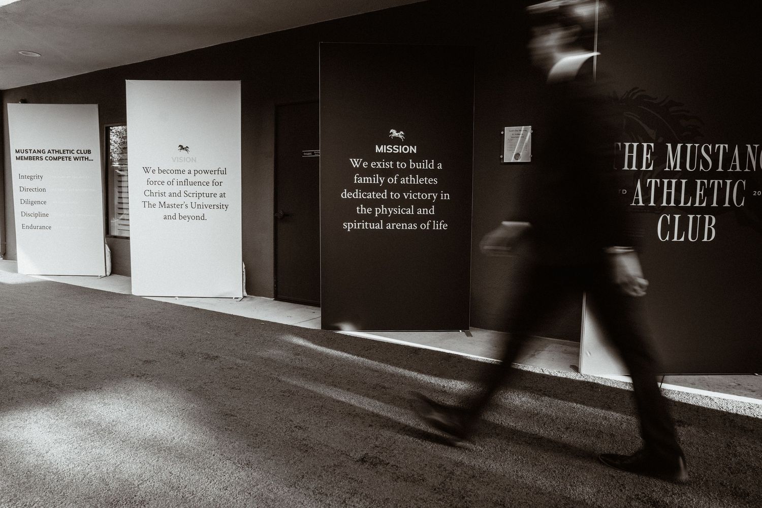 Blurred figure walks past The Mustang Athletic Club motivational wall displays in black and white.