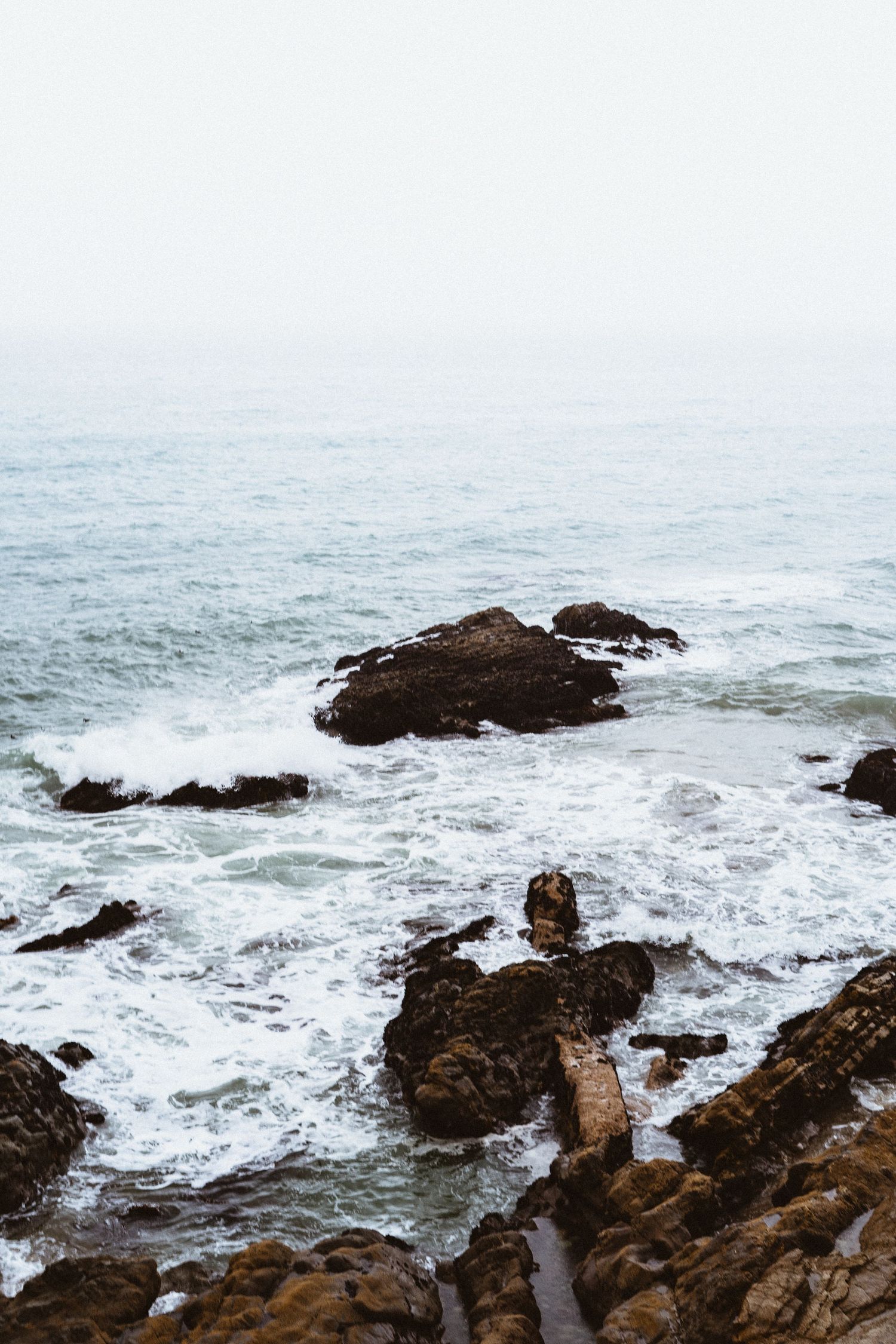 Rocky coastal shoreline with crashing ocean waves under an overcast grey sky.