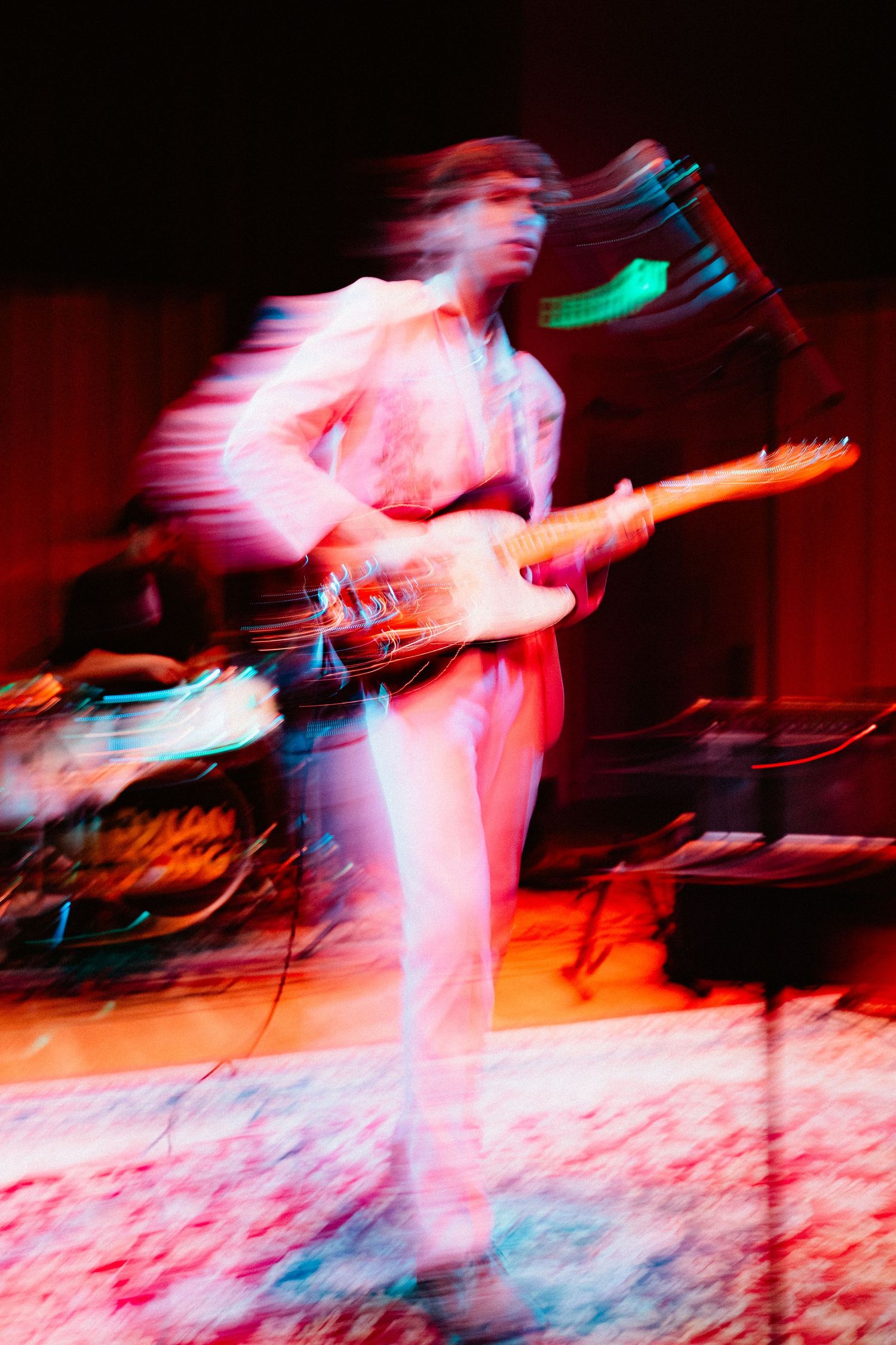 Motion blur photo of guitarist in white outfit playing electric guitar on stage during concert.
