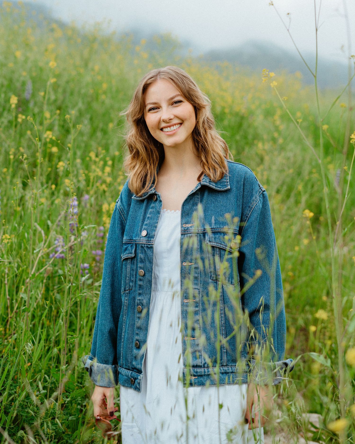 Smiling young woman in denim jacket and white dress stands in a lush wildflower meadow.