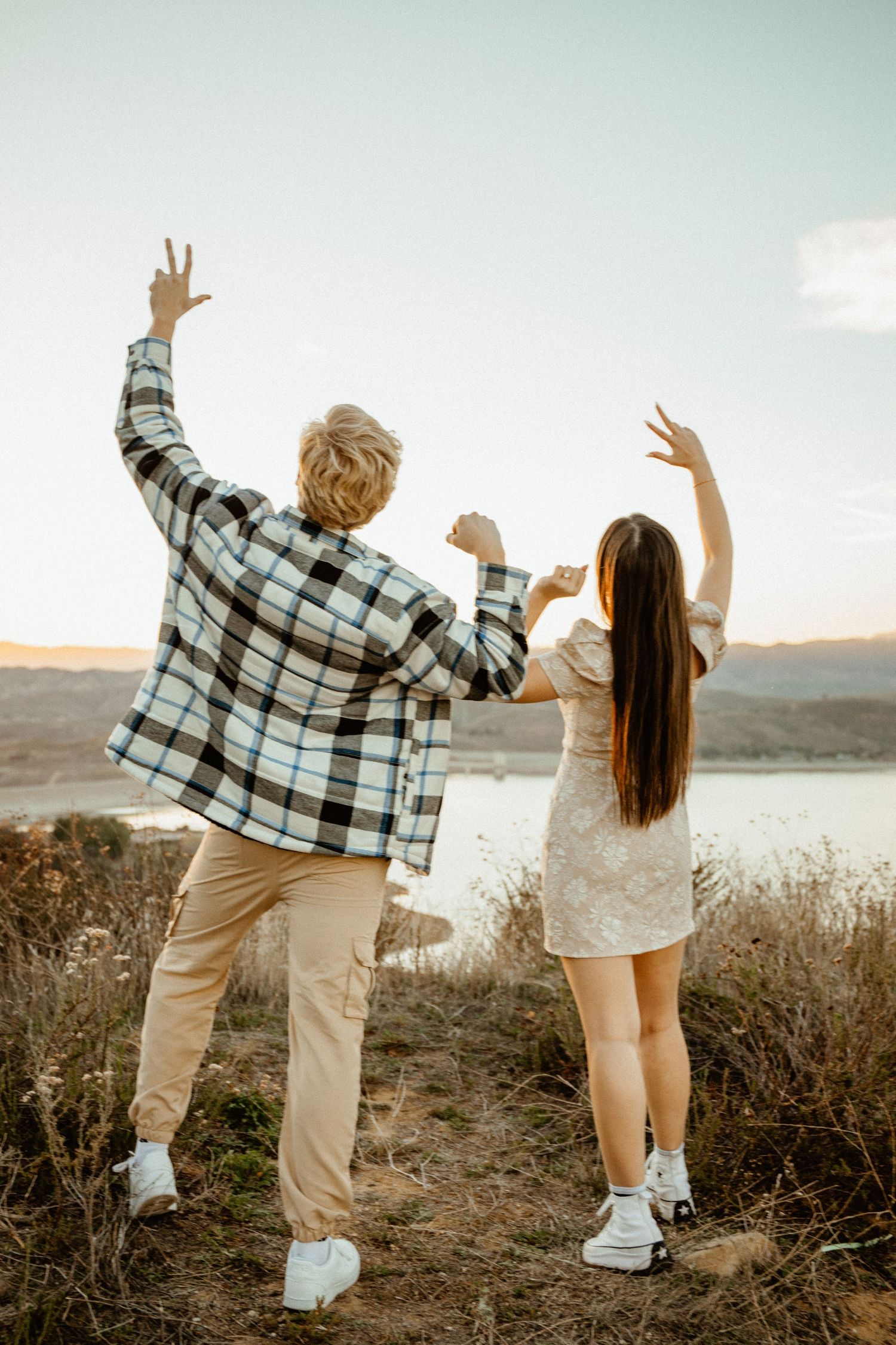 Excited couple celebrating engagement with peace signs overlooking a scenic lake at sunset.