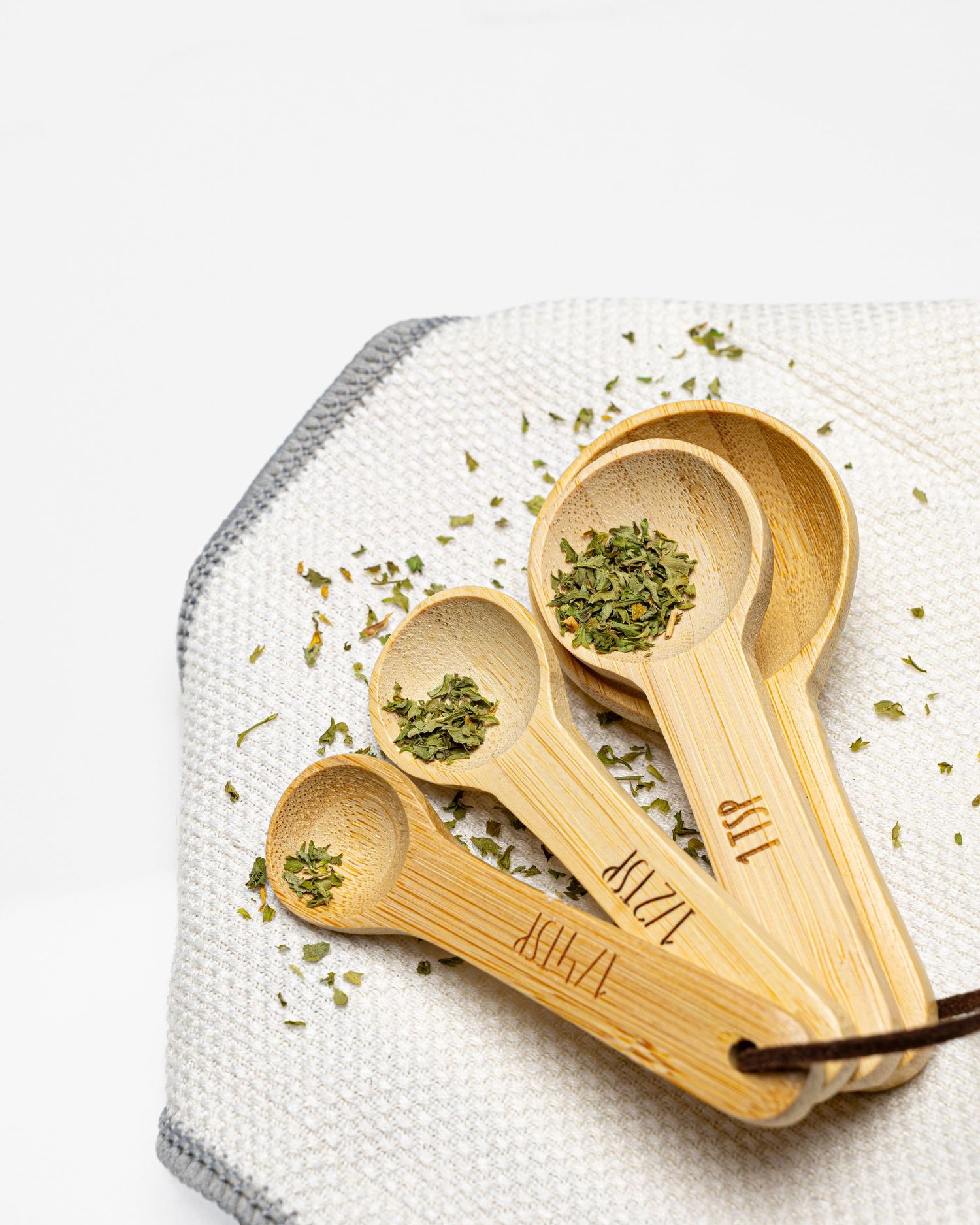 Set of four bamboo measuring spoons with dried herbs on a white textured cloth background.