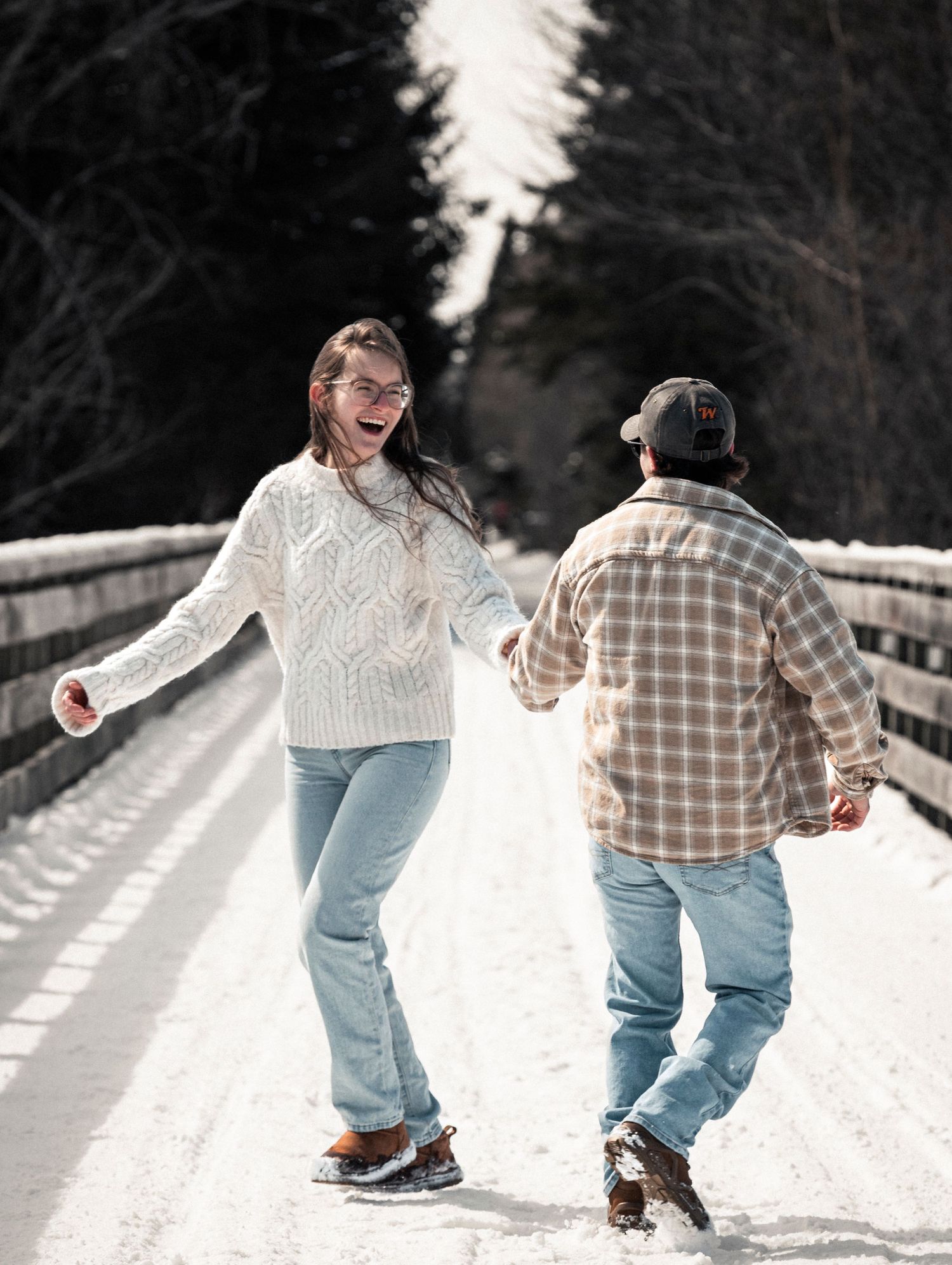 Couple holding hands and dancing playfully on a snowy bridge in winter, wearing jeans and cozy sweaters.