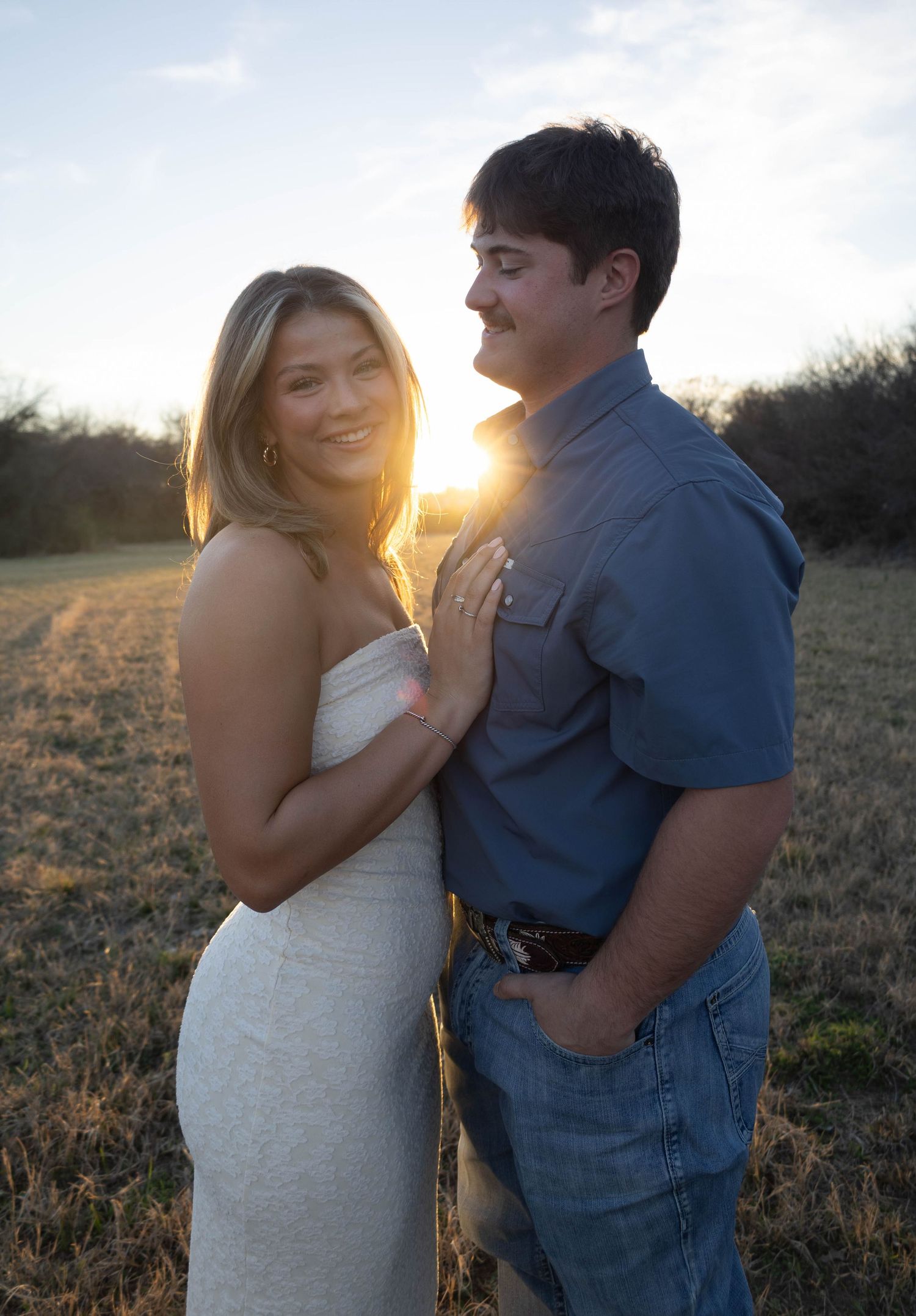 A couple poses romantically at golden hour in a field, woman in white dress smiling as sun sets behind them.