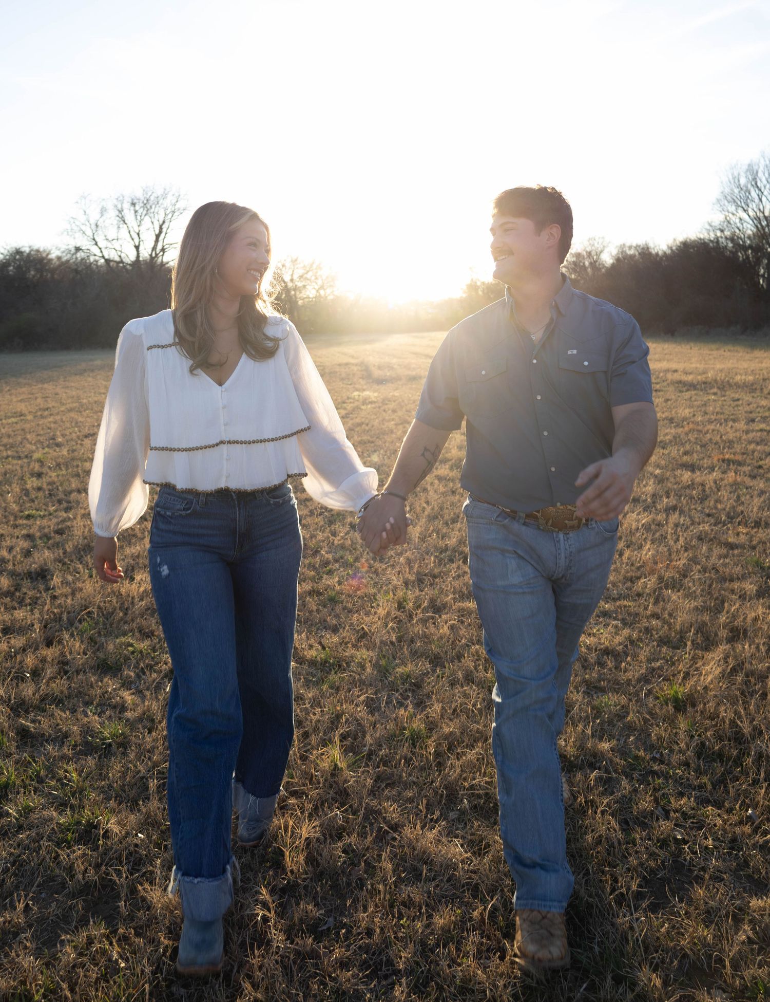 Couple holding hands walking through a sunlit field at golden hour, wearing jeans and casual tops.