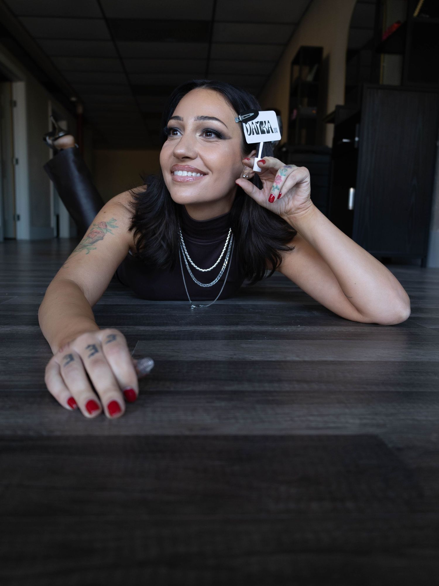 Smiling woman with tattoos lying on a dark wood floor, holding a small sign with red nails and layered necklaces.
