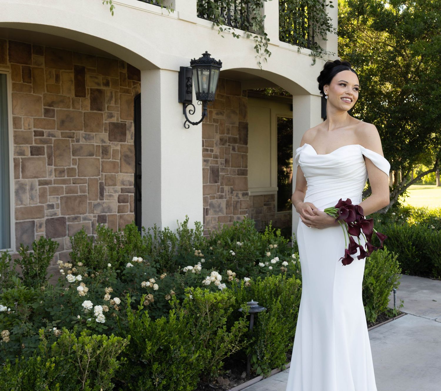 Bride in white off-shoulder gown holding burgundy bouquet stands before stone villa with lush greenery.