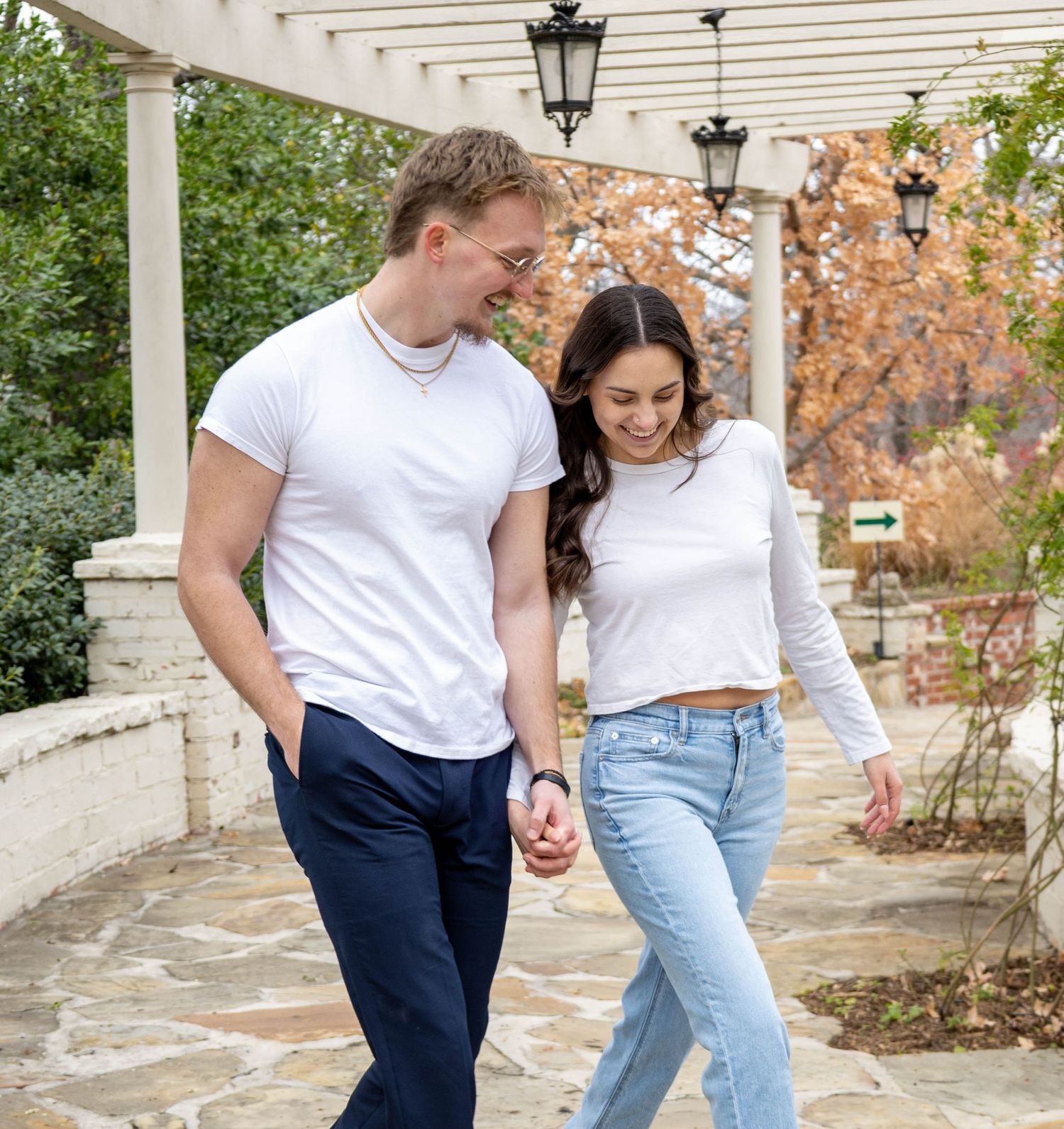 A couple in matching white t-shirts walks together under a covered porch with lanterns and autumn foliage.