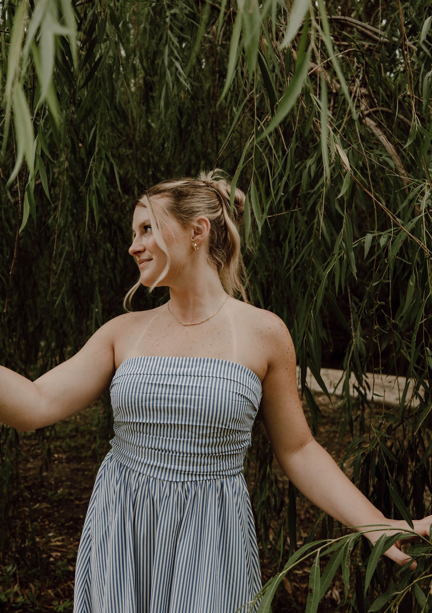 Smiling blonde woman in striped strapless dress standing among weeping willow branches outdoors.