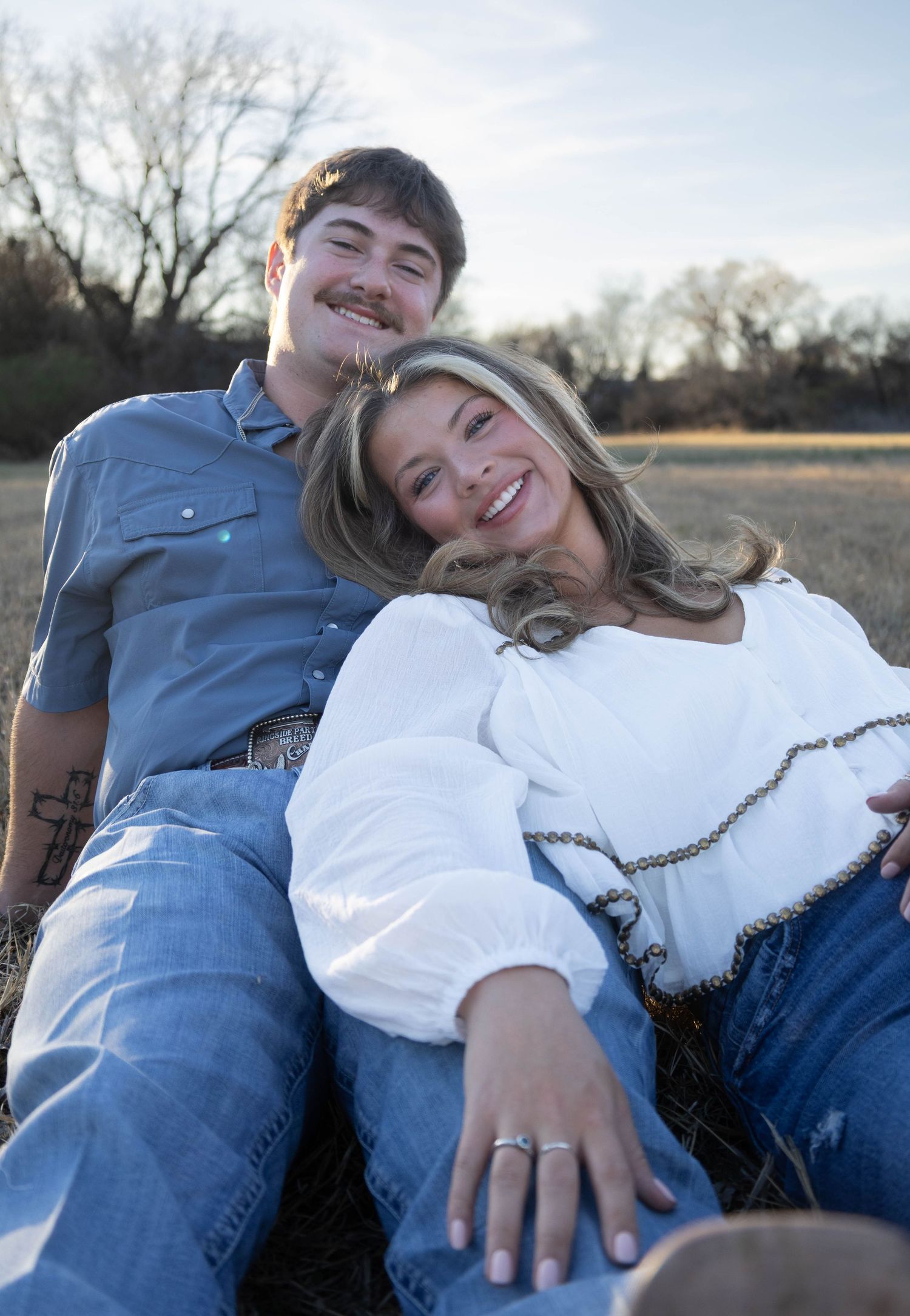 A smiling couple relaxing in a field, wearing jeans and casual shirts, with the woman leaning back on the man.