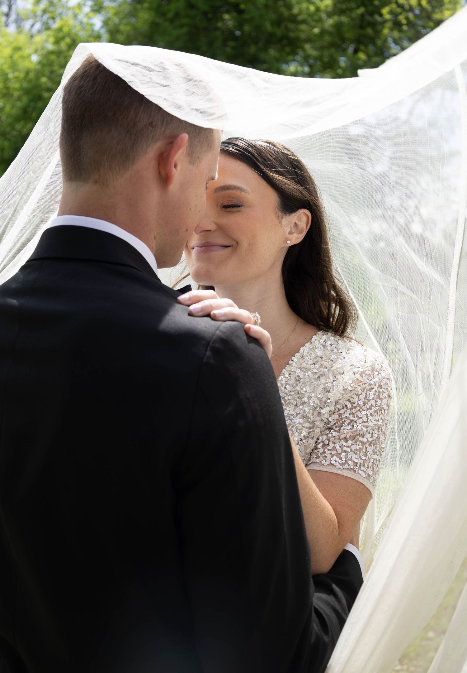 Bride and groom share a romantic moment under a flowing white veil outdoors on their wedding day.