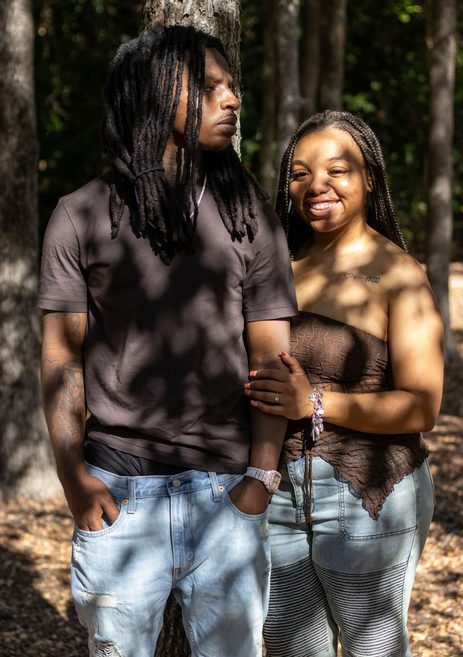 A couple poses together in a wooded area, wearing matching brown tops and light jeans, with dreadlocks hairstyles.
