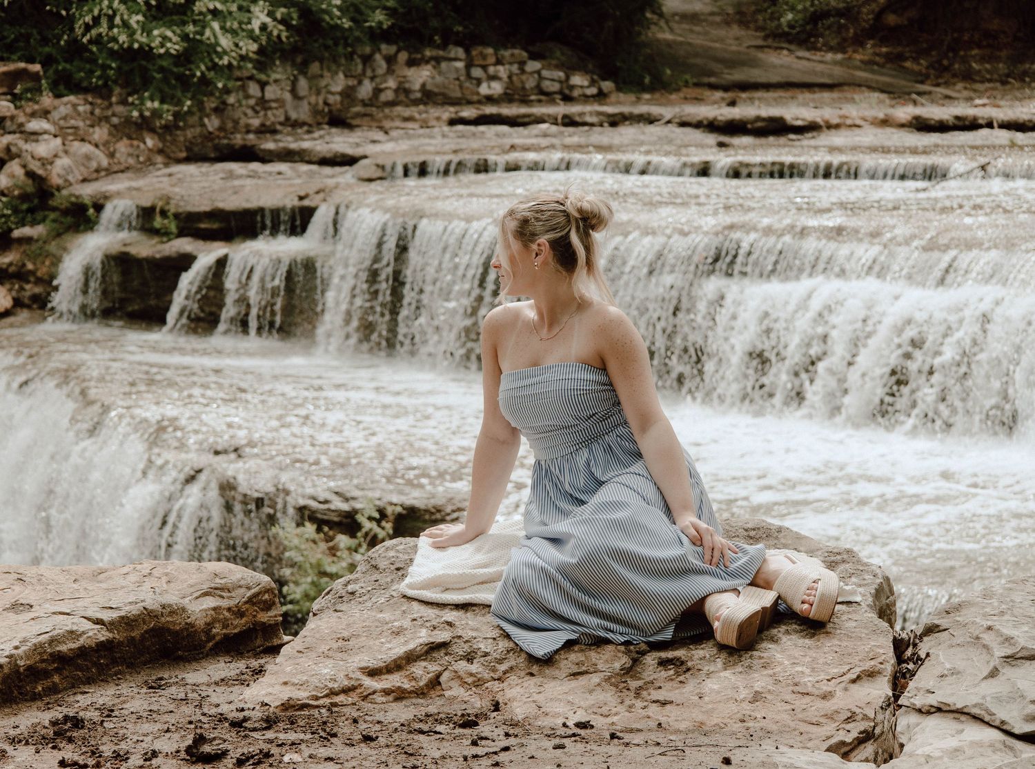 Woman in blue dress sitting on rocks beside a cascading waterfall surrounded by lush greenery.