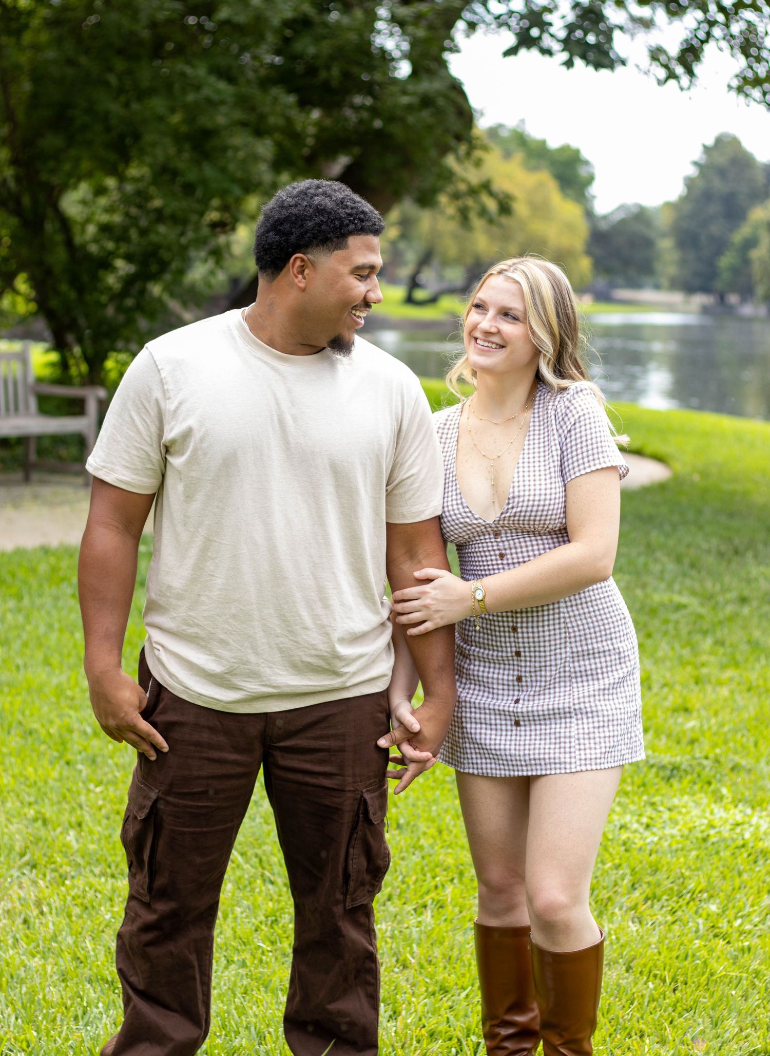 A smiling couple walks hand-in-hand through a lush green park near a pond on a warm day.