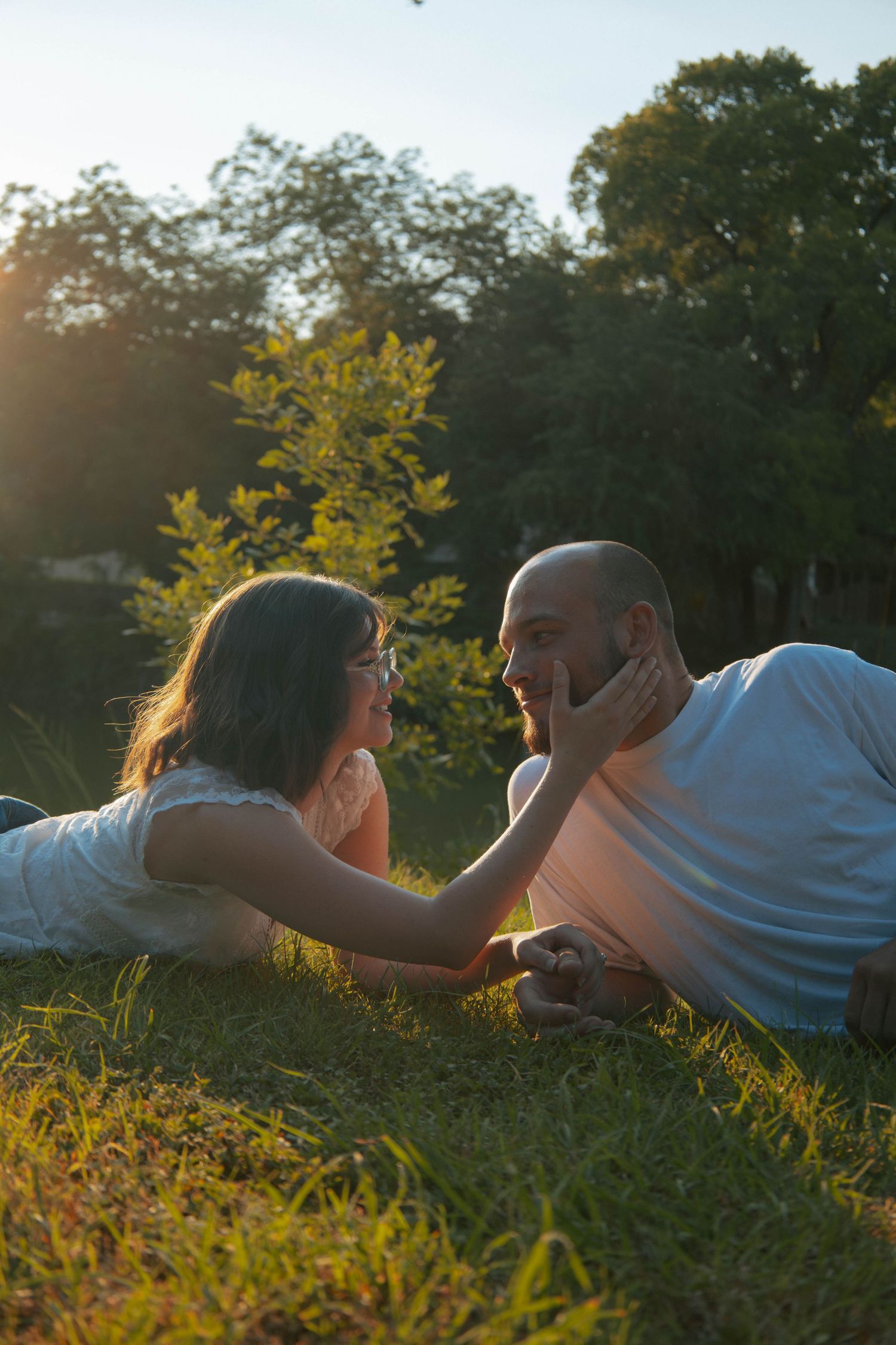 Couple lying on grass in golden hour sunlight, woman touching man's face, sharing a romantic moment in a park.