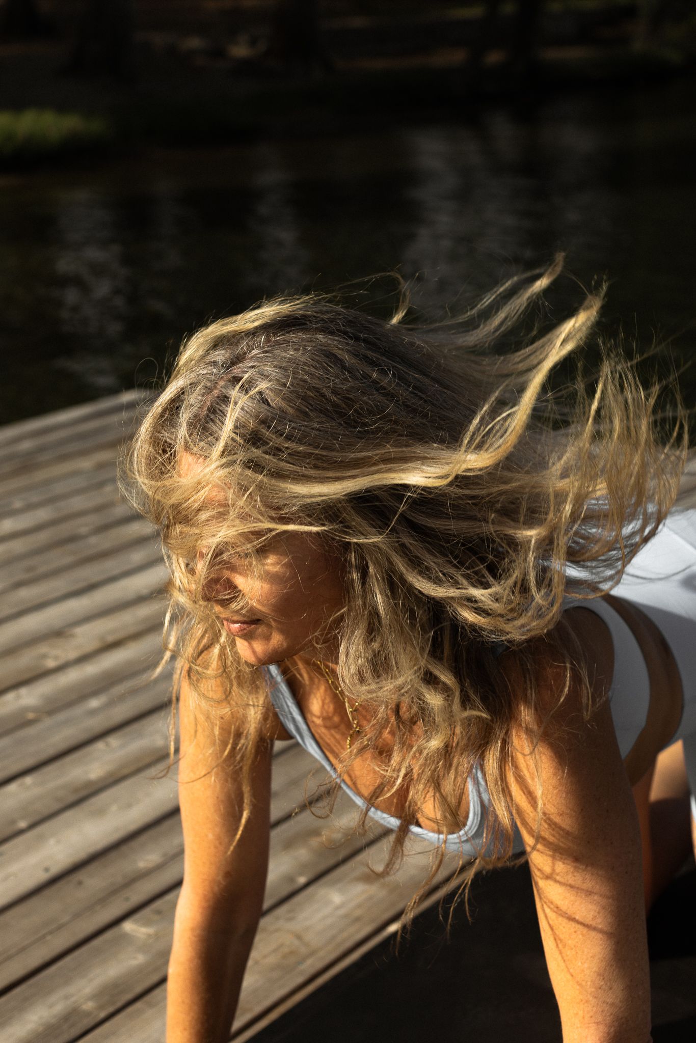 Blonde woman flips her hair forward while exercising on a sunlit wooden dock near a dark lake.