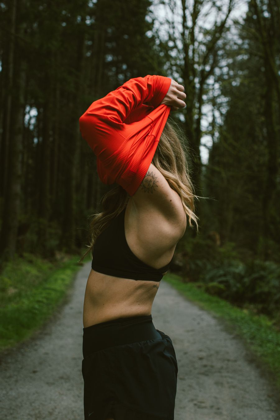 Athlete removing red long-sleeve top on forest trail revealing black sports bra underneath.