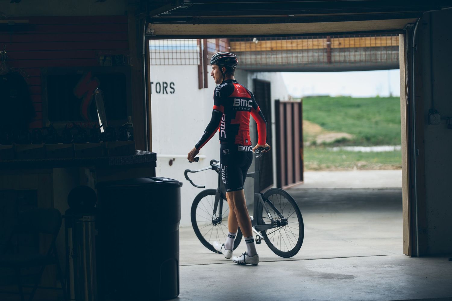 Cyclist in BMC racing kit stands with road bike in garage doorway, ready to ride outdoors.